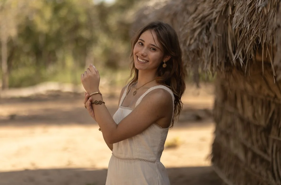 Junge Frau mit braunen Haaren in einem weißen Kleid, lächelnd, steht neben einer Hütte aus Stroh, im Freien bei Sonnenschein.