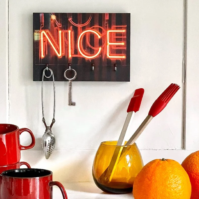 Ein Wandbild mit roter Neonlichtschrift, die 'NICE' sagt, hängt über Küchenutensilien. Darunter befinden sich eine rote Tasse, eine schwarze Tasse, ein gelbliches Glas mit Küchenwerkzeugen, und zwei Orangen.