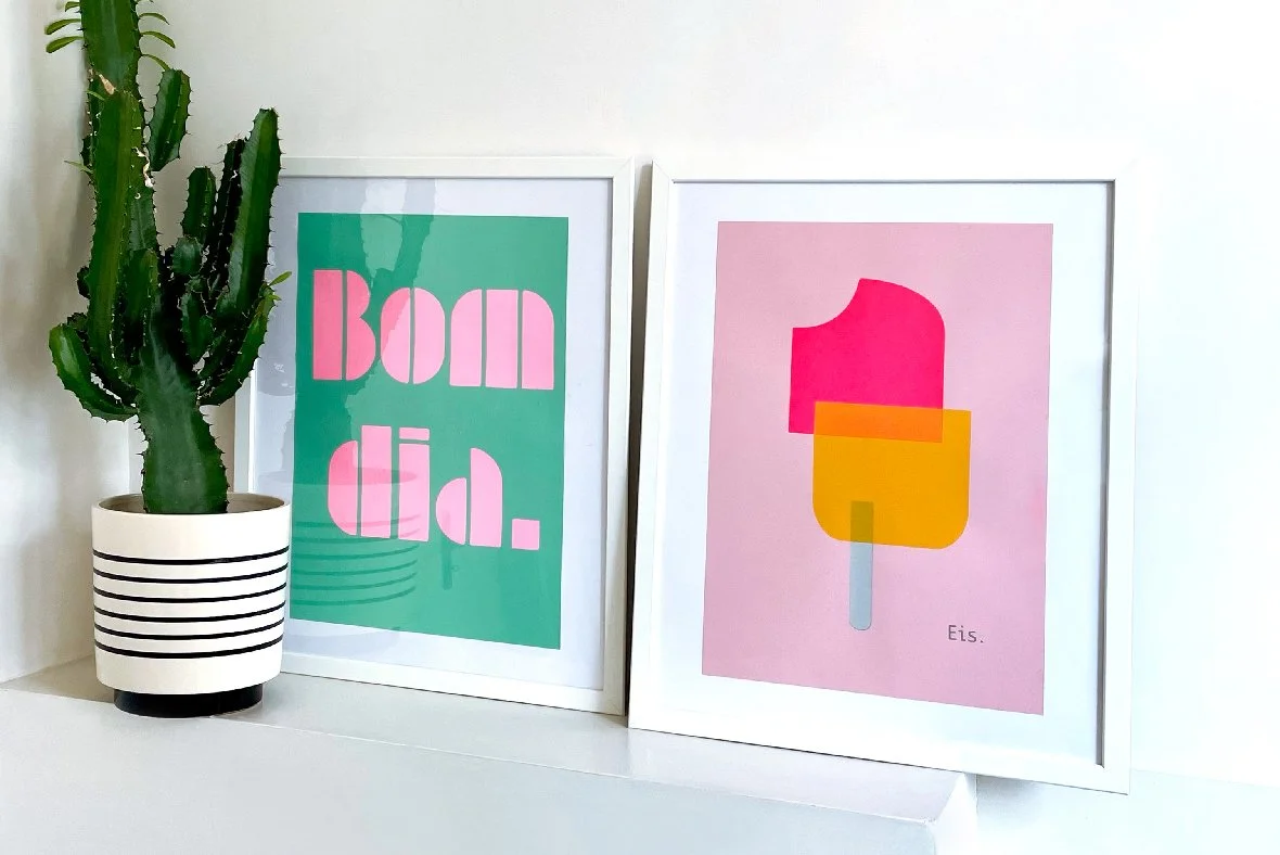 Ein grüne Kaktuspflanze in einer weißen Topfpflanze mit schwarzen Streifen, zwei eingerahmte Poster, eines mit pinken Text "BOM DIA." auf grünem Hintergrund, das andere mit einer Illustration eines bunten Eis am Stiel auf rosa Hintergrund, darunter steht "Eis."