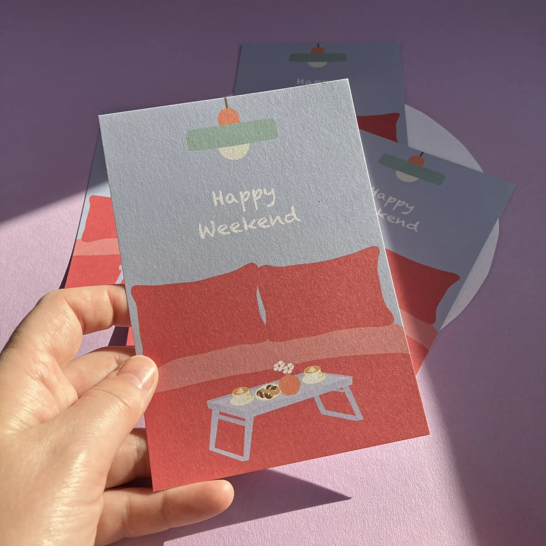 Grußkarte mit einem roten Sofa, einer Couchtisch mit Kerzen, Keksen und Blumen, und der Text "Happy Weekend" auf einem hellblauen Hintergrund, mit weiteren Karten im Hintergrund