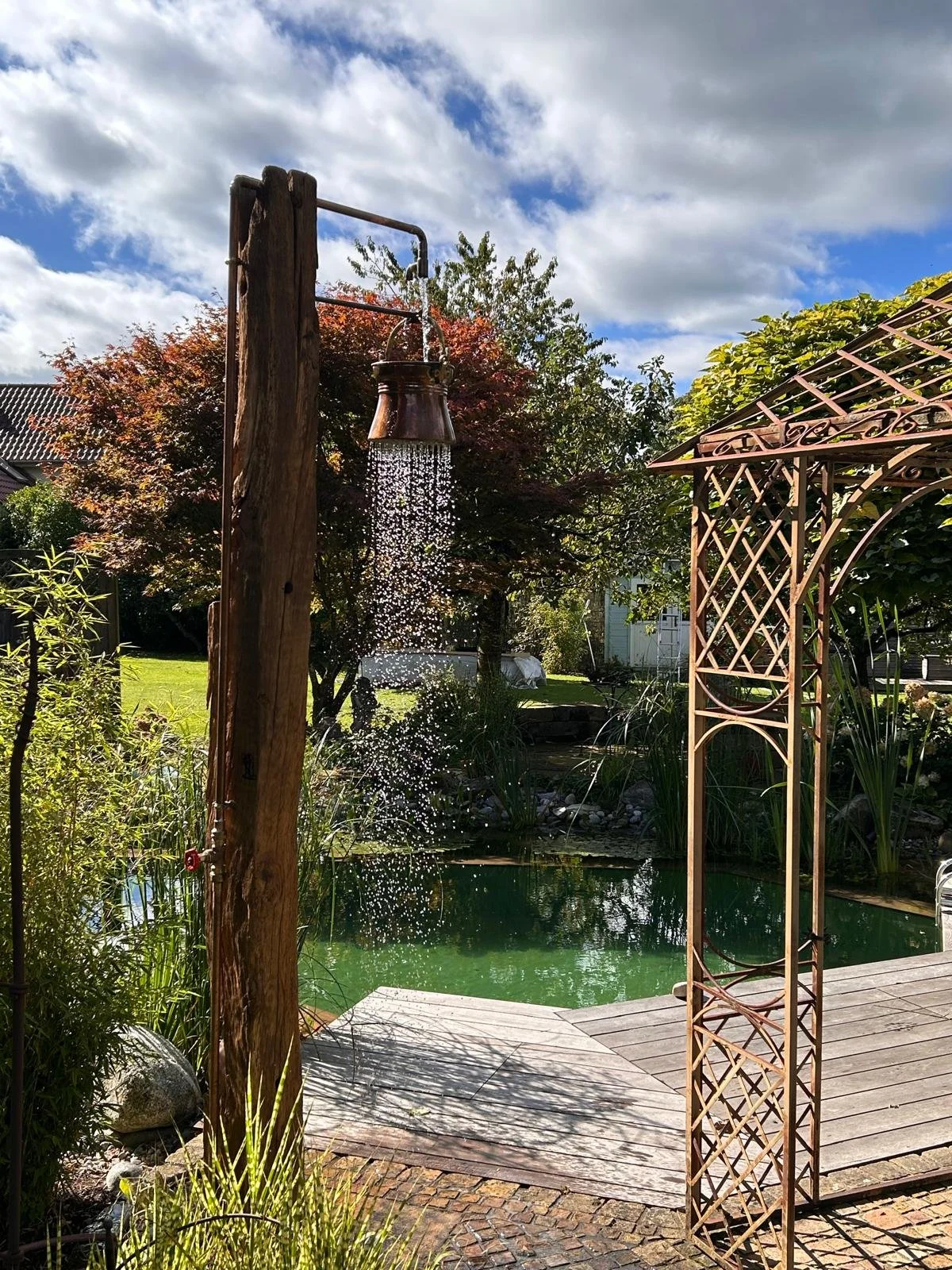 Ein Garten im Sonnenlicht mit einem kleinen Teich, einem Baum, einer Holzdusche zum Gießen und einem filigranen, rustikalen Metallbogen.