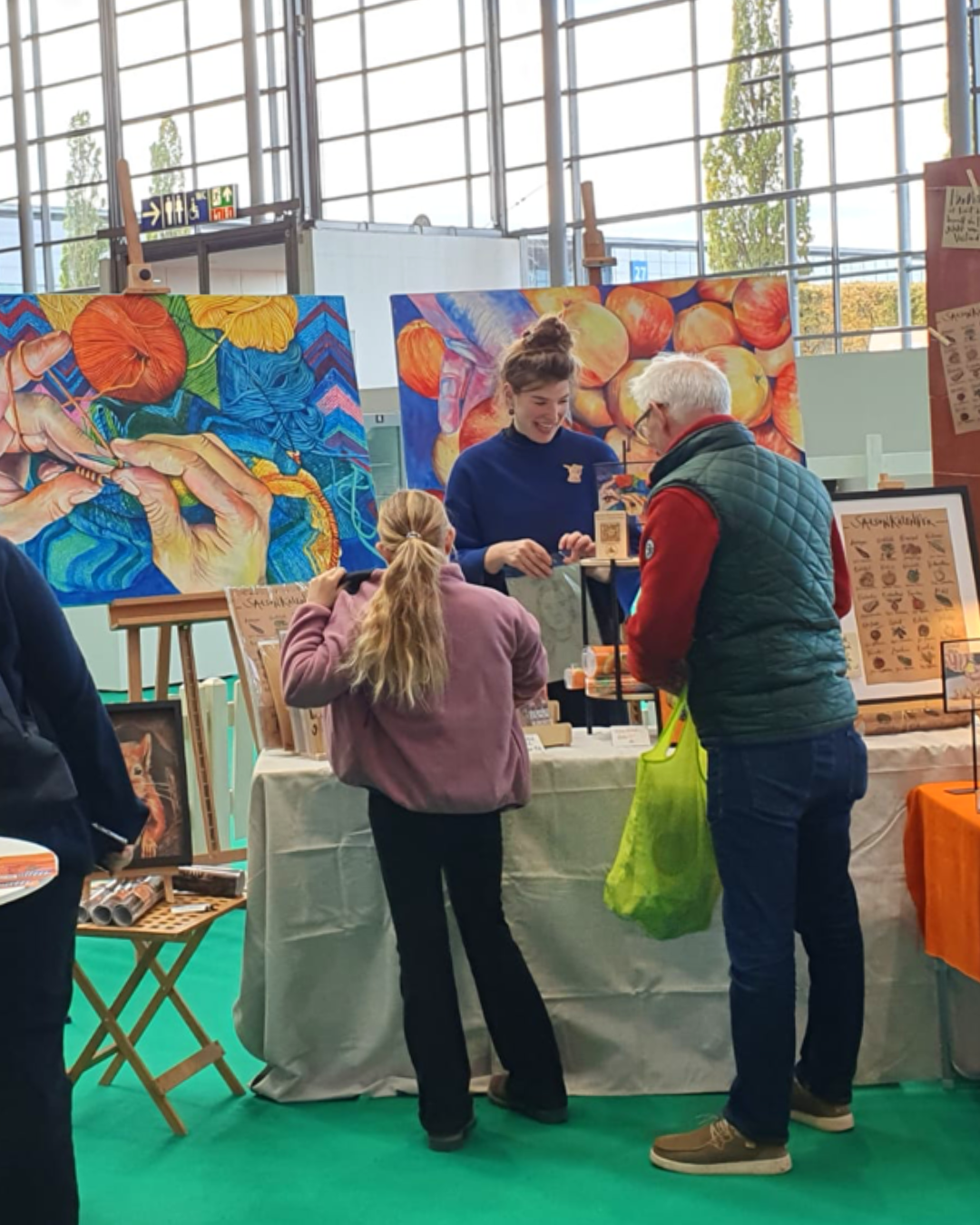 Menschen an einem Messestand, der Kunstwerke und Handwerkswaren verkauft, im Hintergrund bunte Gemälde von Pflanzen und Früchten.