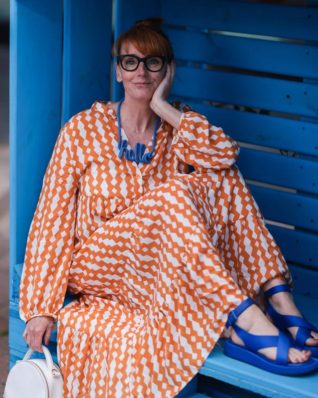 Eine rot haarkleidete Frau mit Brille in einem orange-weißen Kleid und blauen Sandalen sitzt auf einer blauen Bank, die in einer blue Painted Struktur ist. Sie trägt eine Halskette mit dem Wort 'hello' und hält eine weiße Tasche.