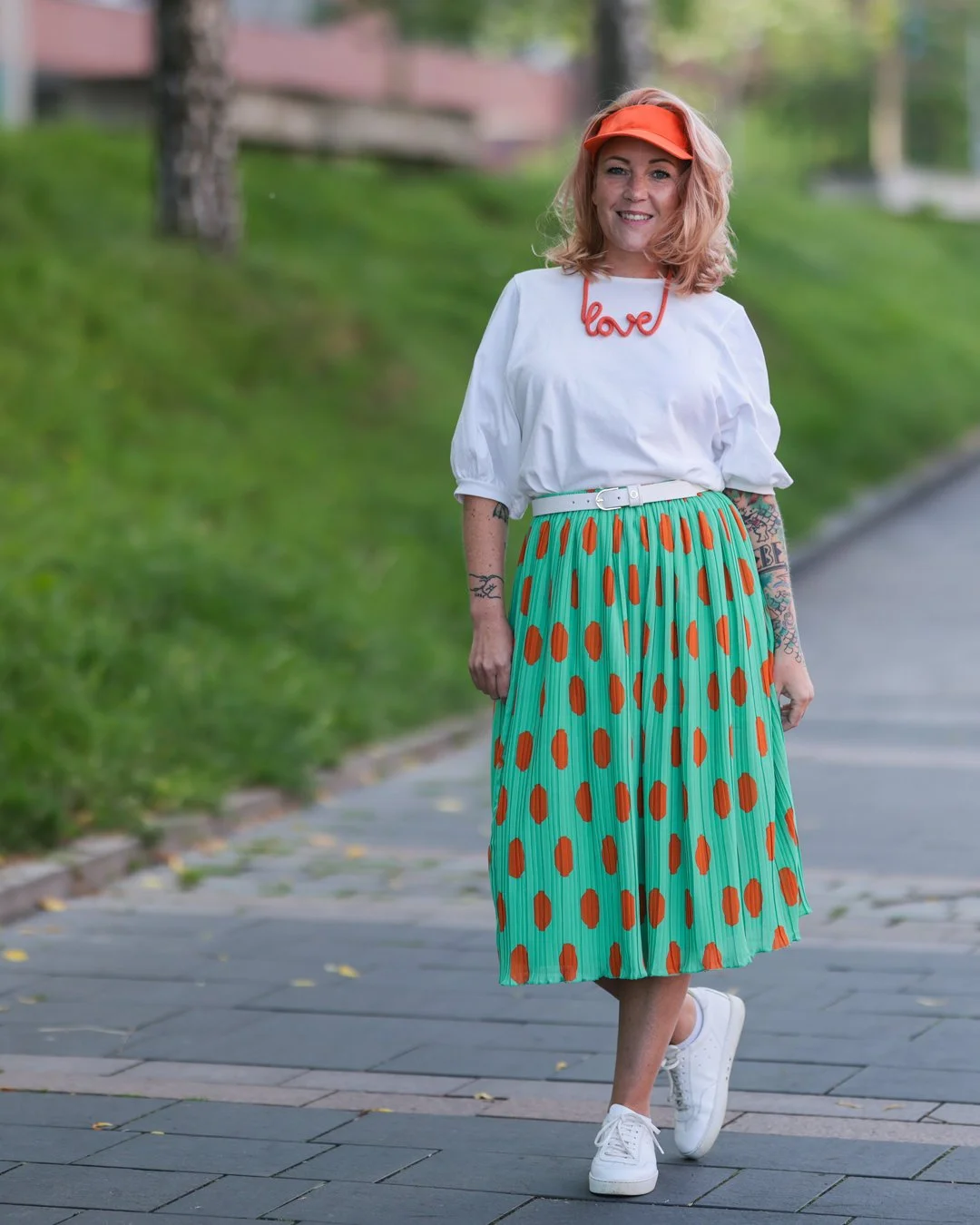Frau mit Schulter-Tattoo, weißen Schuhen, bunter Wickelrock mit orange Punkten, weißem T-Shirt mit "love"-Schriftzug, orange Cap, Spaziergang auf Gehweg in parkartiger Umgebung.