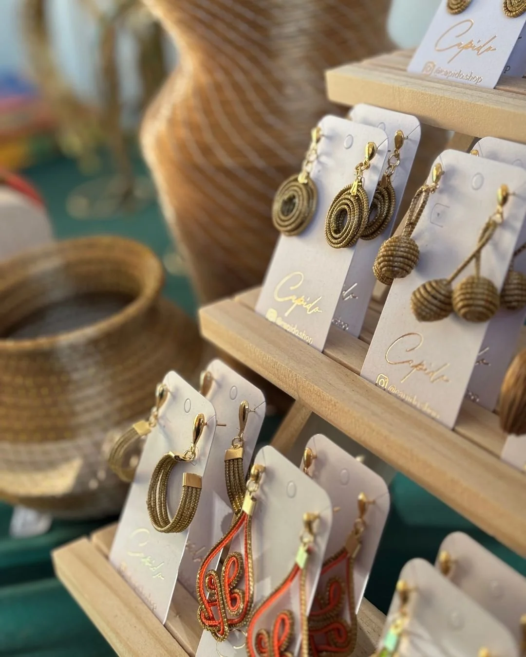 Verschiedene goldene Schmuckstücke, darunter Ohrringe, auf einer Verkaufsklemmbrett mit dem Label 'Capula', in einem Verkaufsstand aus Holz. Zusätzlich sind geflochtene Körbe im Hintergrund sichtbar.