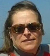 Una donna con capelli castani raccolti, occhiali da sole scuri e un vestito rosa, con sfondo blu.