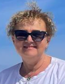 Donna anziana con capelli ricci e corti, occhiali da sole neri, sorriso, indossa una maglietta bianca, sfondo chiaro