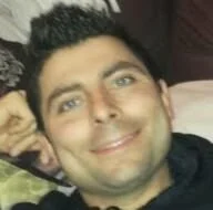 Prima piano di un uomo sorridente con capelli scuri e occhiali, che si trova in un ambiente interno.