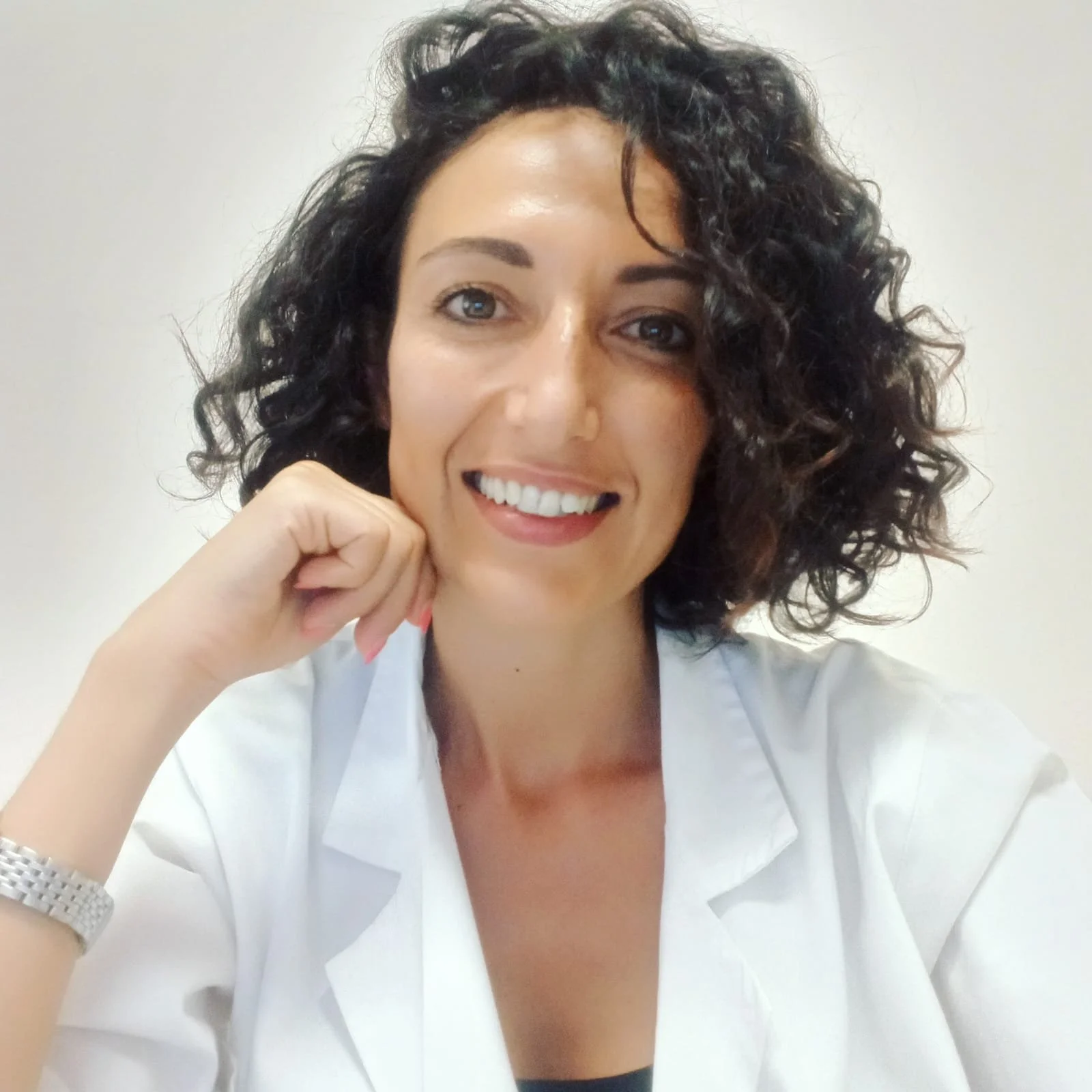 Donna sorridente con capelli ricci, indossa una camicia bianca, riposa il volto sulla mano, sfondo neutro.