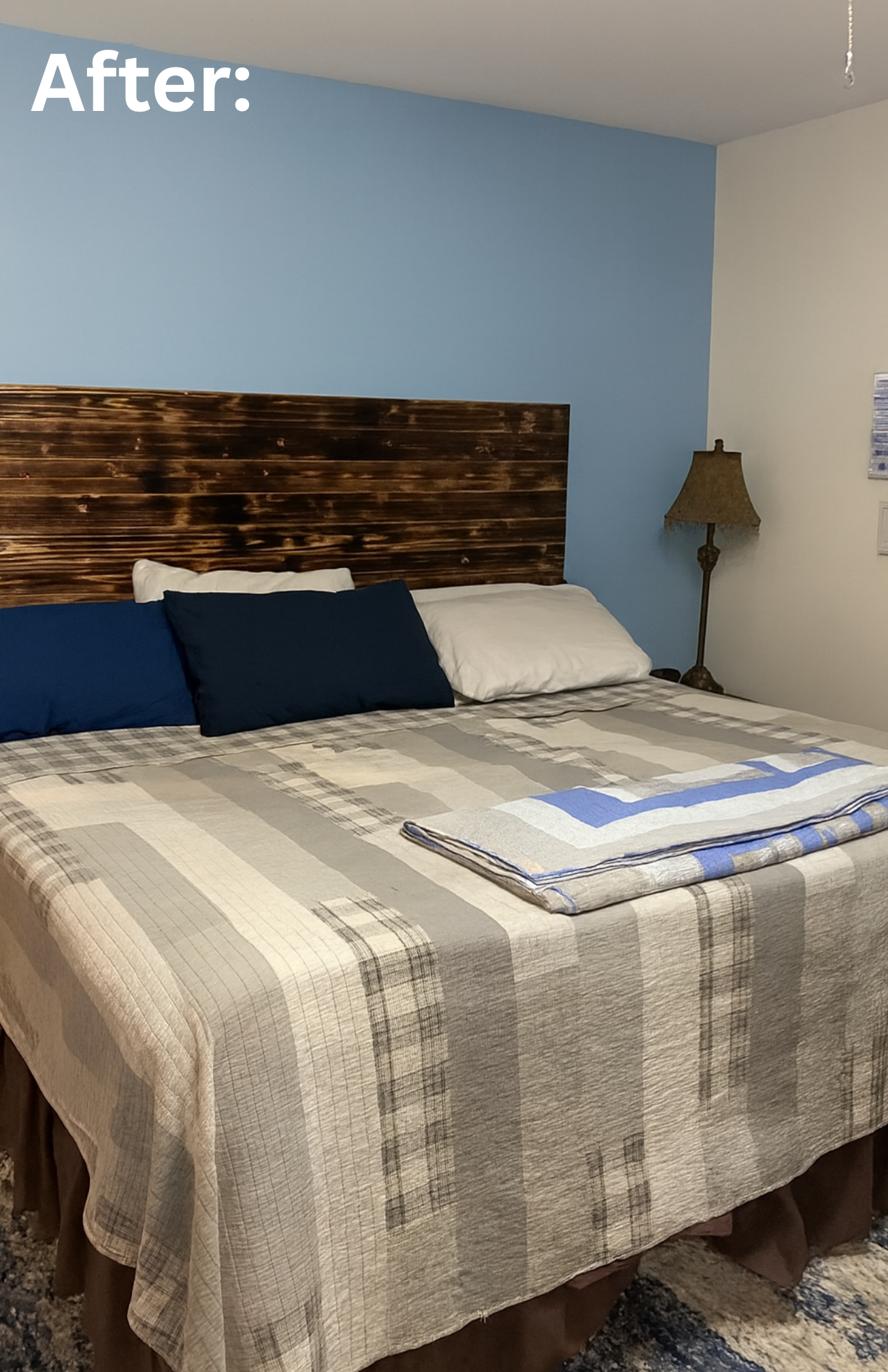 A neatly made bed with a plaid beige and gray bedspread, and a burned wood headboard, in a bedroom with a light blue and off-white wall and a table lamp beside the bed. Showcasing the after photo of repainting this bedroom.
