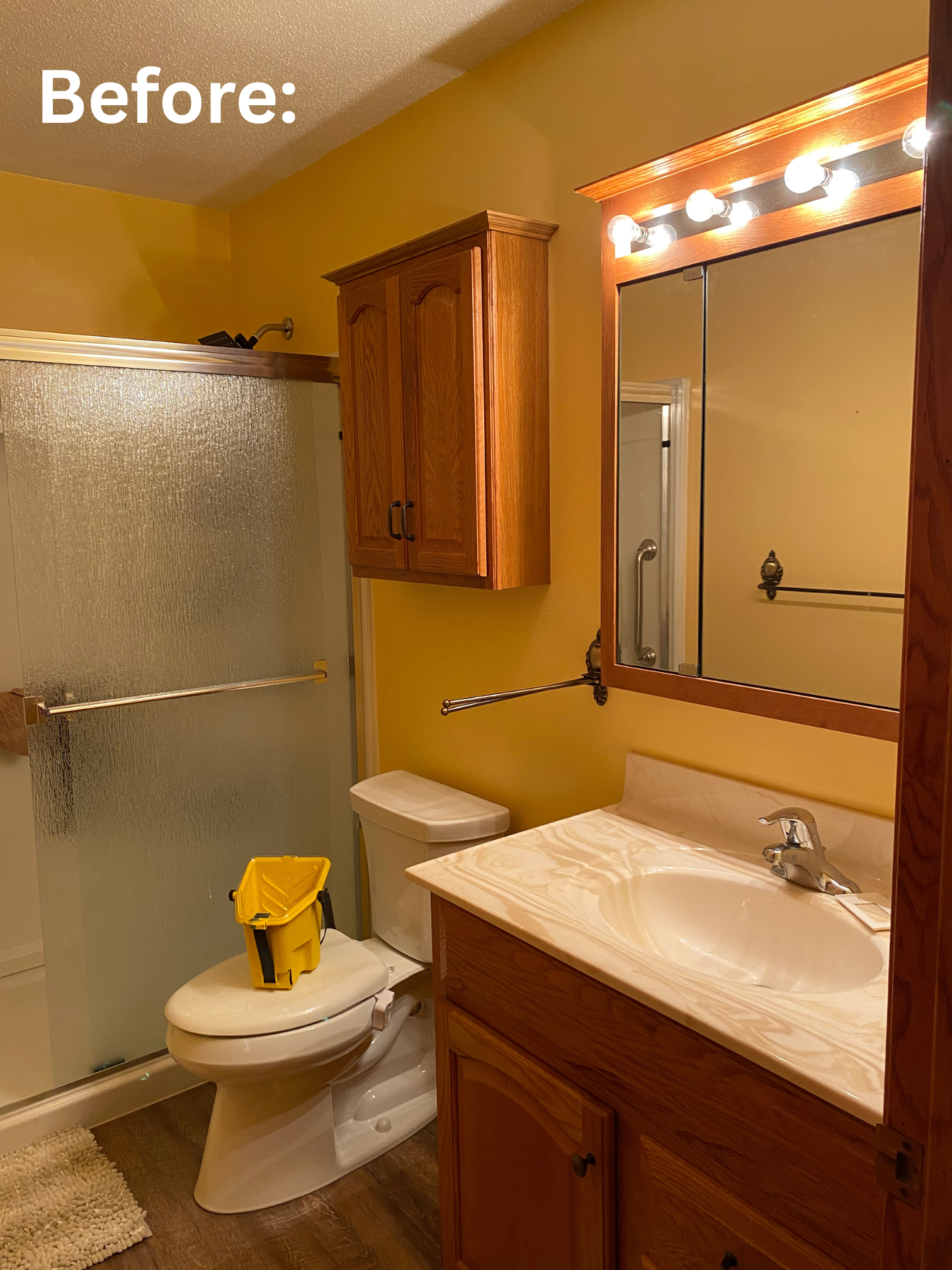 Bathroom with yellow walls, wooden cabinets, a mirror with lights, a toilet, a sink with a marble countertop, a shower with a frosted glass door, and a beige rug on the dark wood floor. Showcasing the before photo of repainting this bathroom.