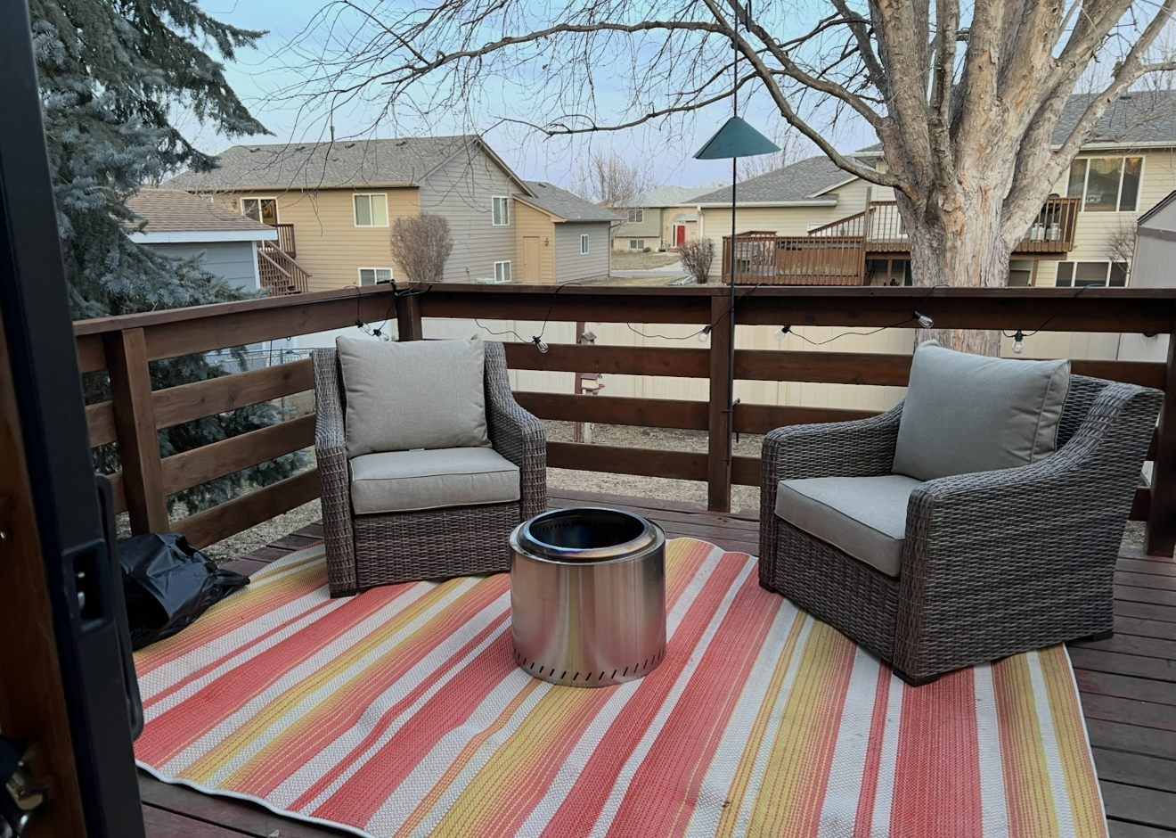 Outdoor wooden deck with two wicker chairs and a colorful striped outdoor rug, with a metal fire pit between the chairs, overlooking neighboring houses and trees.