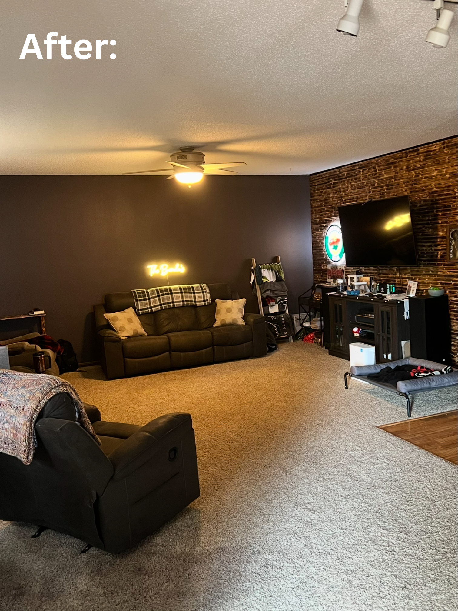 Living room with a ceiling fan, dark accent wall with neon sign, leather couch, TV on a burned wood accent wall, small dog bed, and various decorations and furniture. Showcasing the after photo of the newly painted living room.
