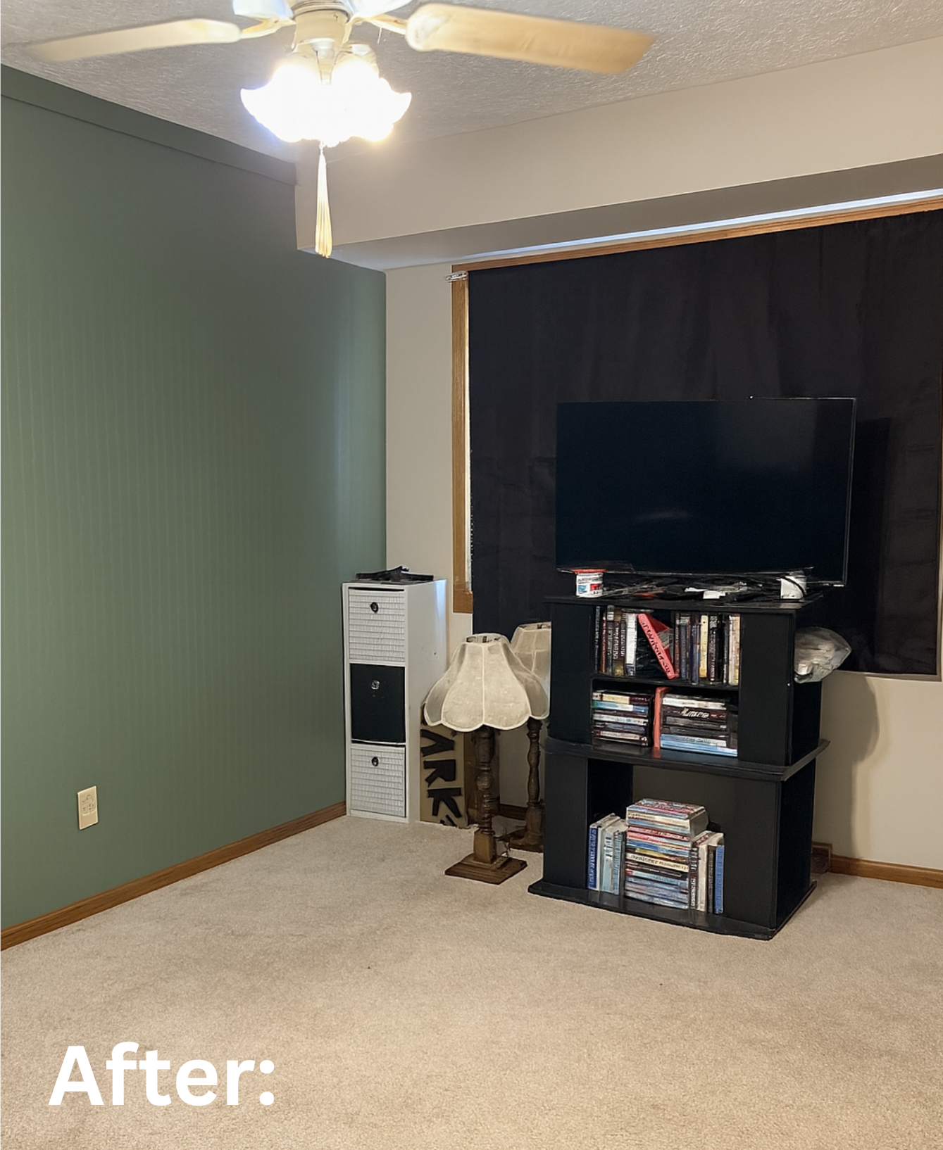 A living room corner with a ceiling fan, green accent wall, black entertainment center with TV, books, and DVDs, a small wooden side table with a lamp, and carpeted floor. Showcasing the after photo of the repainted room.
