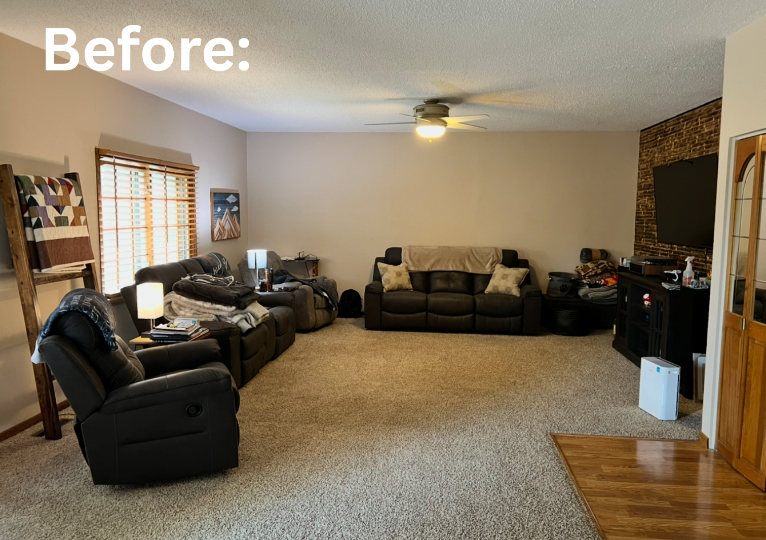Living room with beige carpet, beige walls, and a white ceiling. Contains black and brown sofas, a recliner, window with blinds, wall art, and an entertainment area with a TV on a brick accent wall. There are lamps, a small table, and a fan in the room.