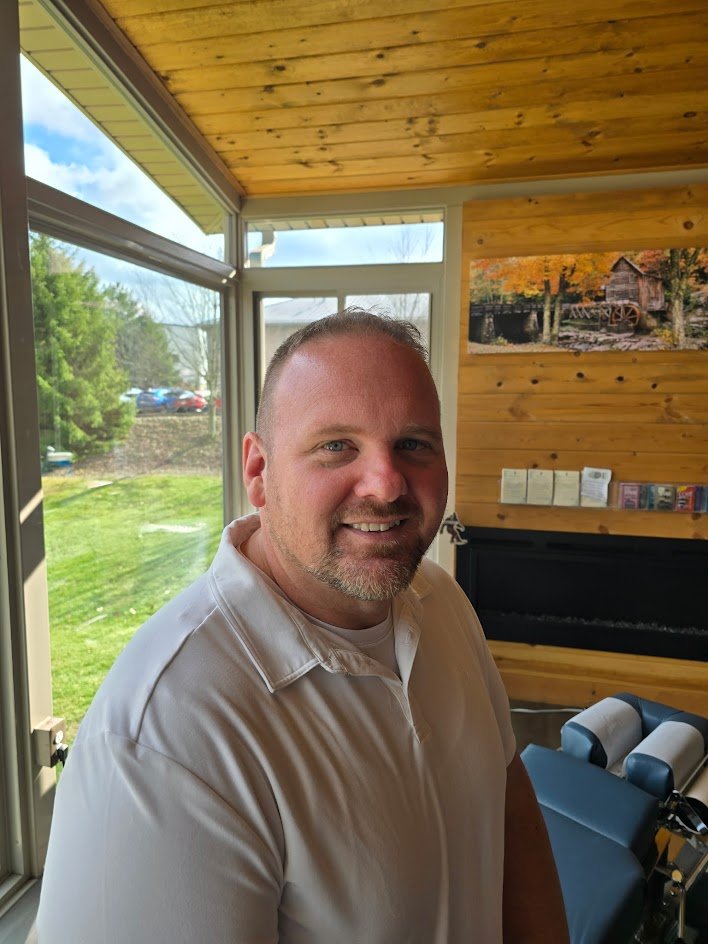 A man with short hair and a beard smiling indoors near large windows with a view of a grassy yard and trees outside.