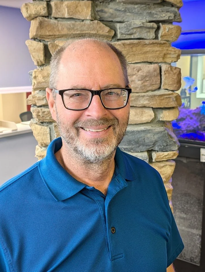 A smiling middle-aged man with glasses, a beard, and short hair, wearing a blue polo shirt, standing in front of a stone wall with an aquarium in the background.