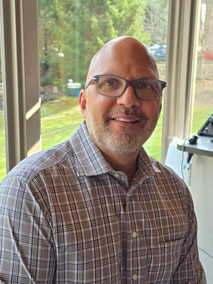 A middle-aged man with glasses, a bald head, and a beard, smiling while sitting indoors near a large window with a view of greenery outside, wearing a plaid button-up shirt.