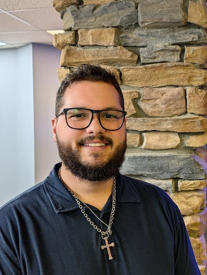 A young man with glasses and a beard smiling in front of a stacked stone wall.