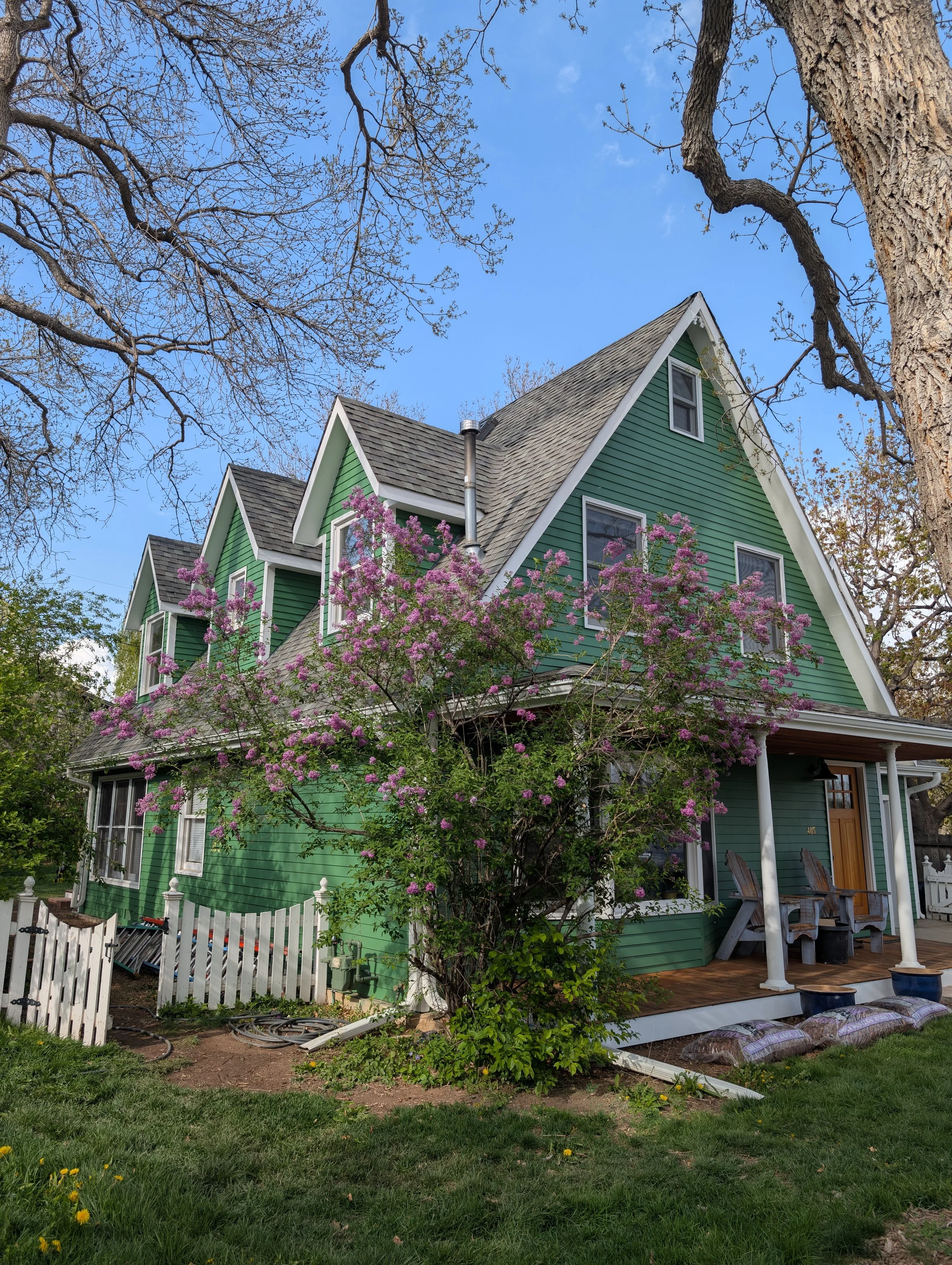 A green, multi-story house with a front porch area featuring wooden chairs. Pink flowering shrub in front, leafless trees around, and a clear blue sky in the background.
