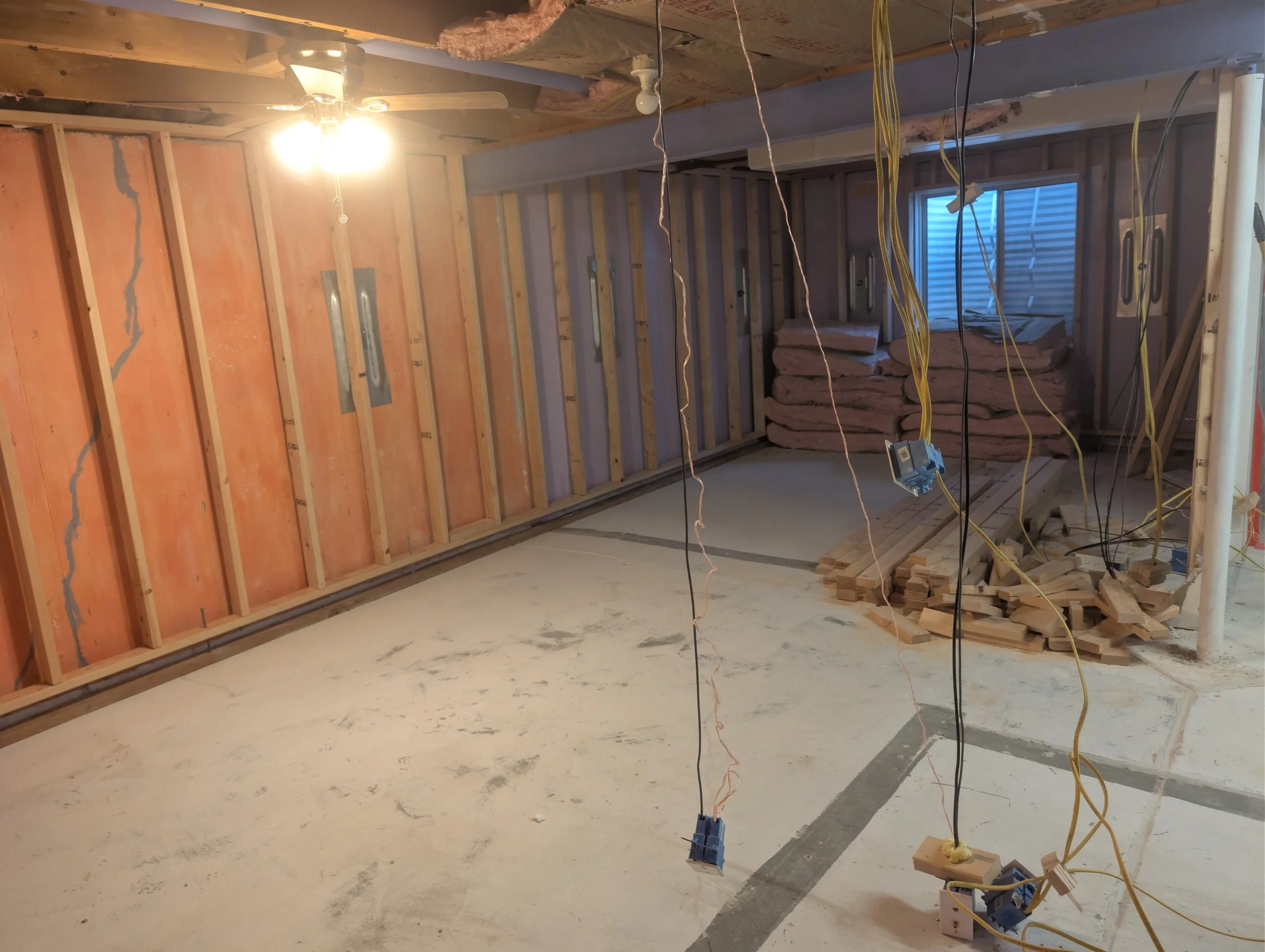 Interior of a building under construction, with exposed wooden framing, electrical wiring hanging, insulation stacked in the corner, and a ceiling fan installed, illuminated by a bright light.