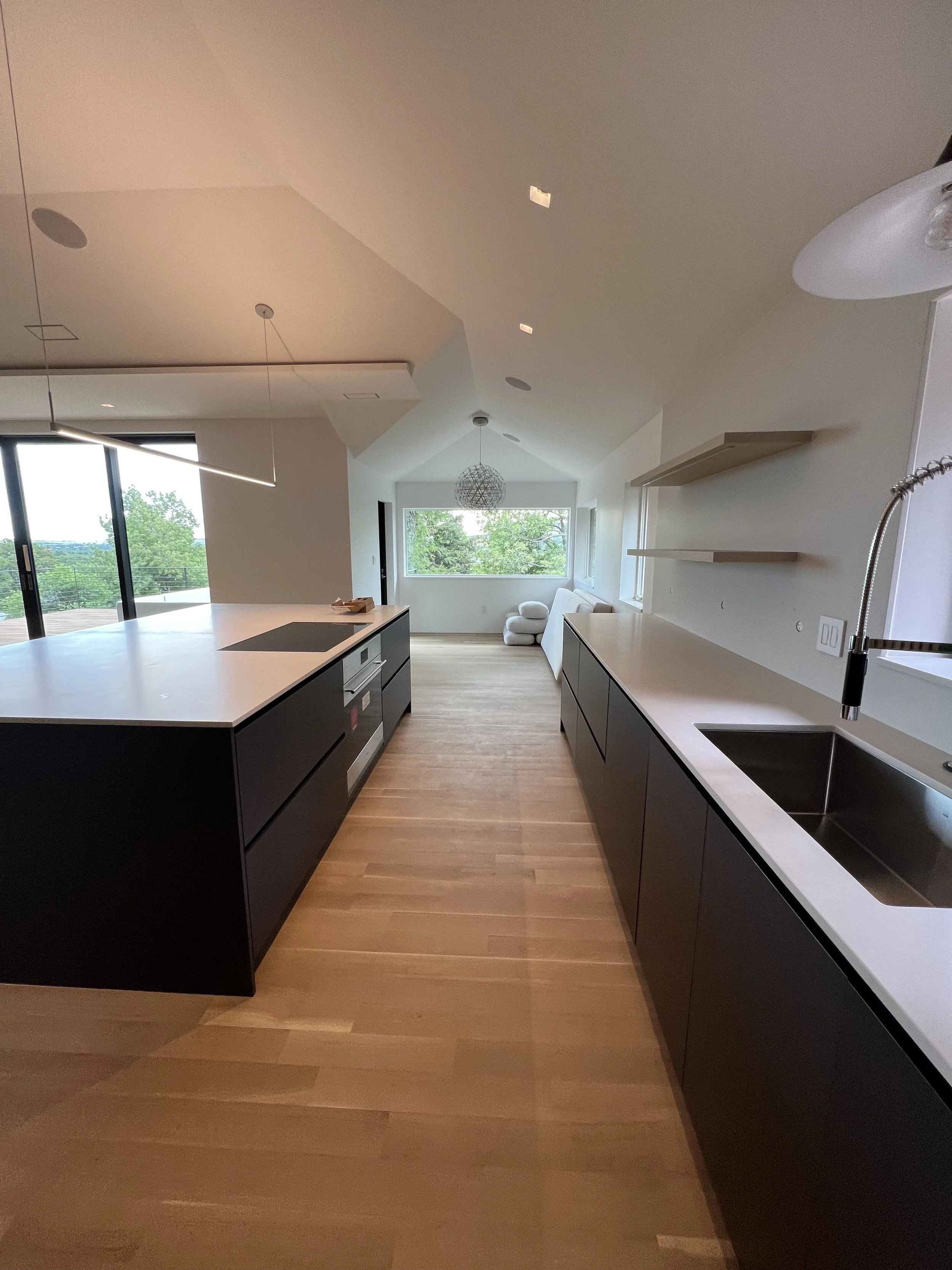 Modern kitchen with black cabinets, white countertops, a sink, hardwood floors, and large windows overlooking greenery, with a distant view of trees outside.