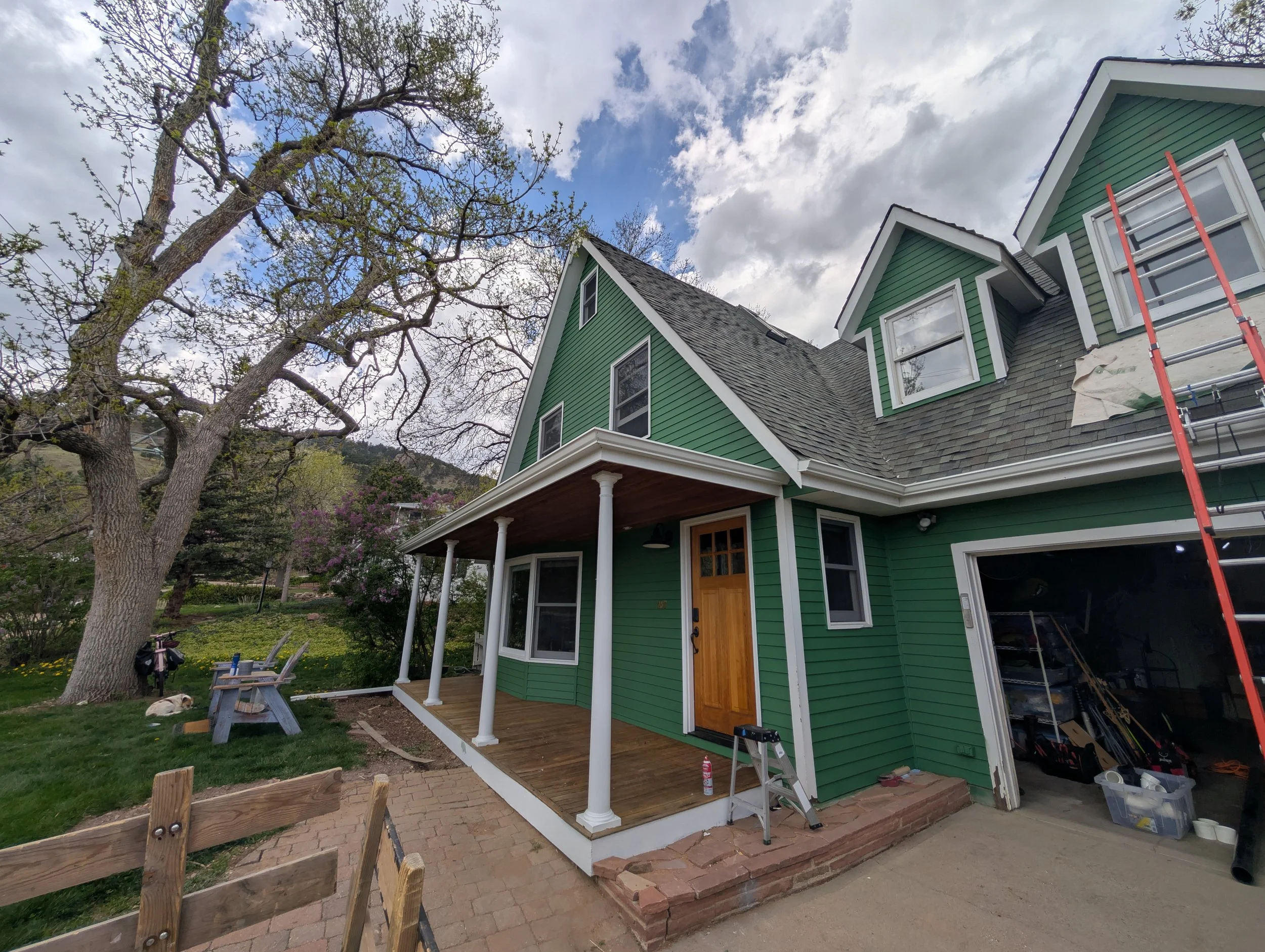 Green house with a front porch and a ladder outside, under a partly cloudy sky, surrounded by trees and a garden with a bench and a dog.