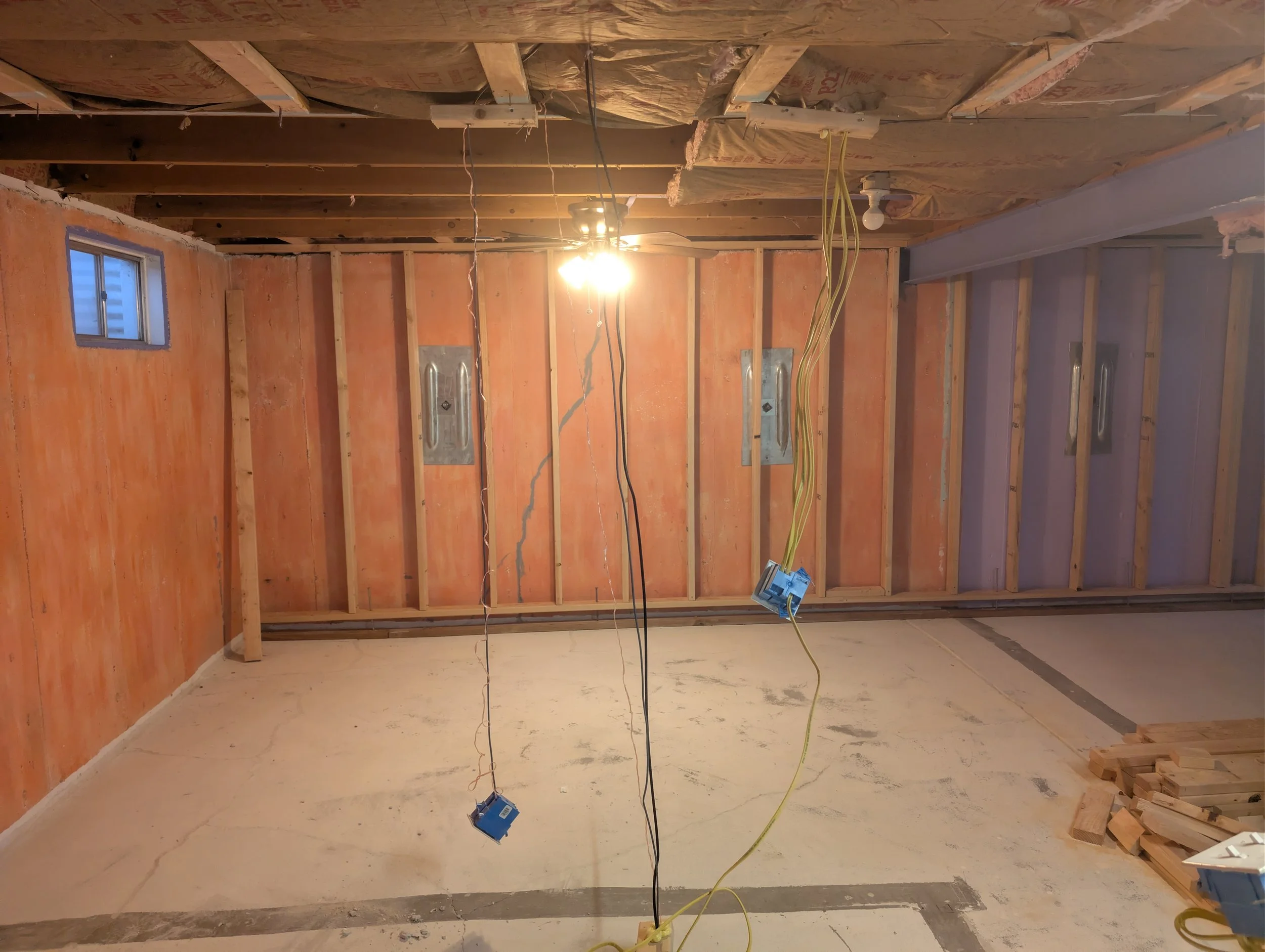 Interior of a room under construction with exposed wooden framing, electrical wiring hanging from the ceiling, and a single bulb hanging from the ceiling providing light.