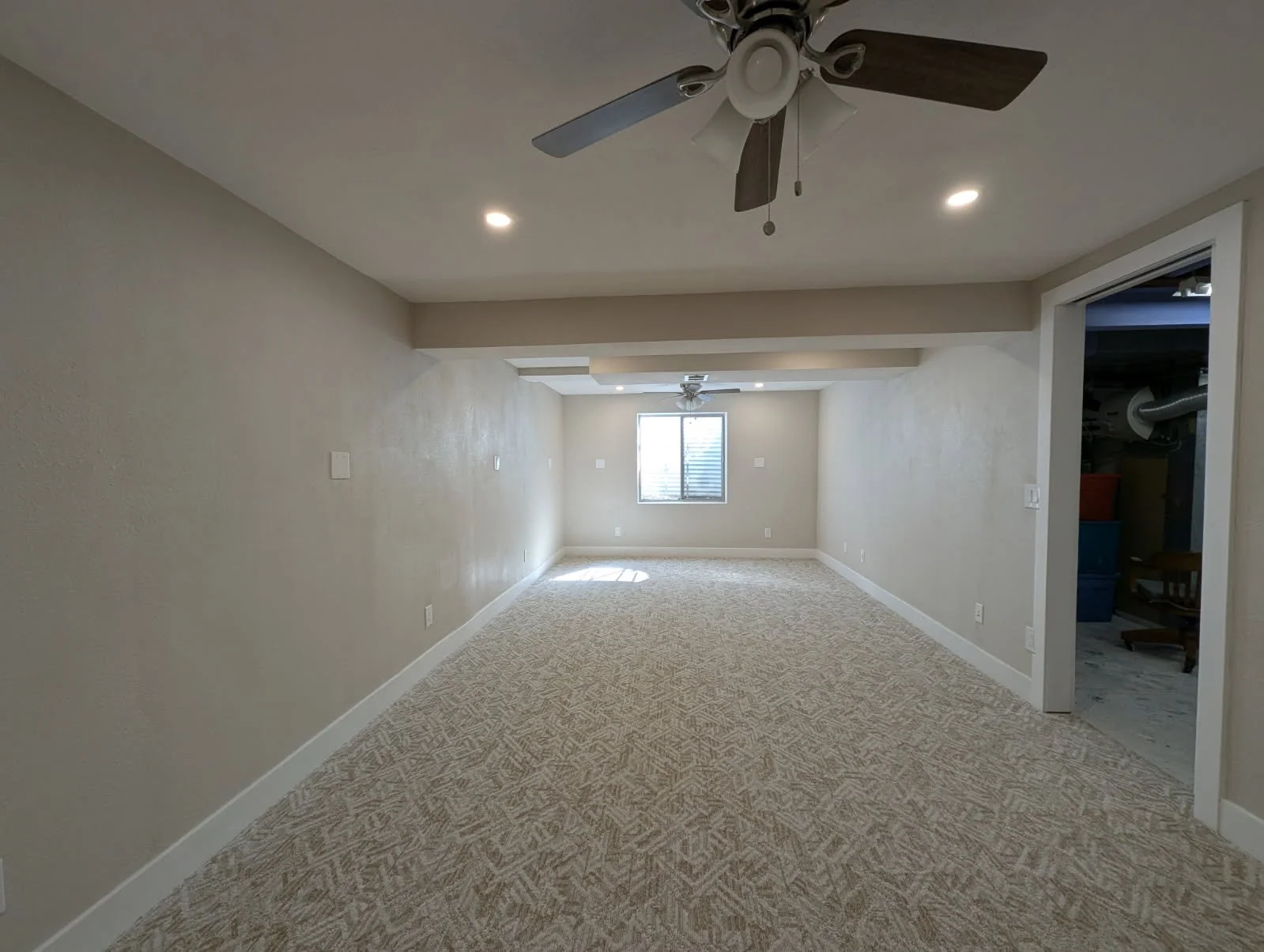 Empty living room with beige walls, beige patterned carpet, ceiling fans, and recessed lighting. There is a window at the far end of the room with a small room or closet on the right.