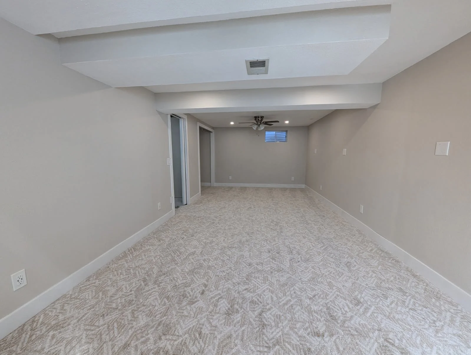 Empty living room with beige carpet, white walls, ceiling fan, small high window, and sliding doors.