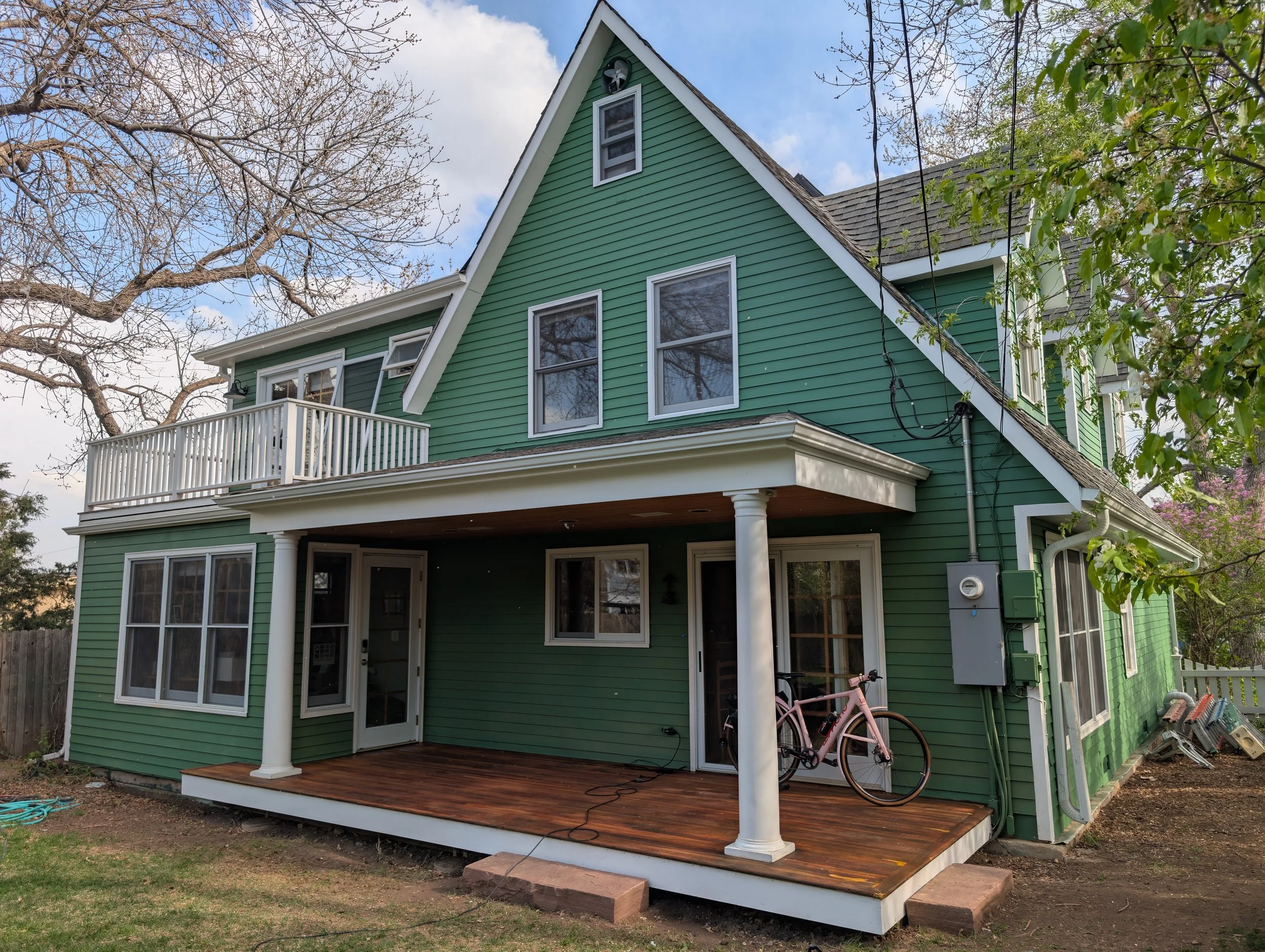 Green two-story house with a wooden porch, bicycle, and white balcony, surrounded by trees.