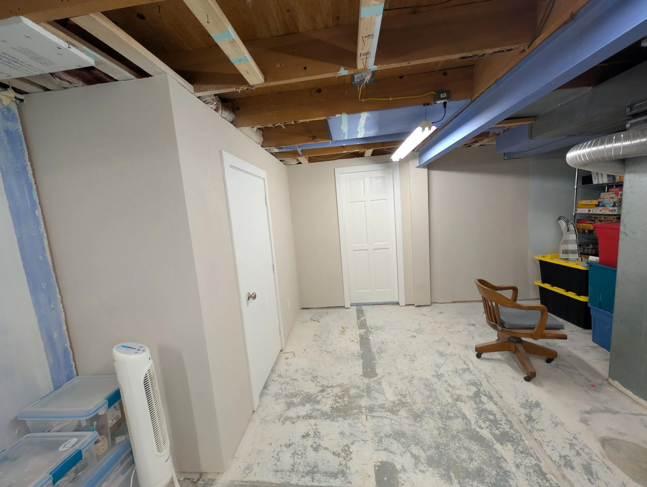 Room under construction or renovation with drywall, exposed wood beams, and storage shelves. There is a white door, a chair, and a fan in the space.