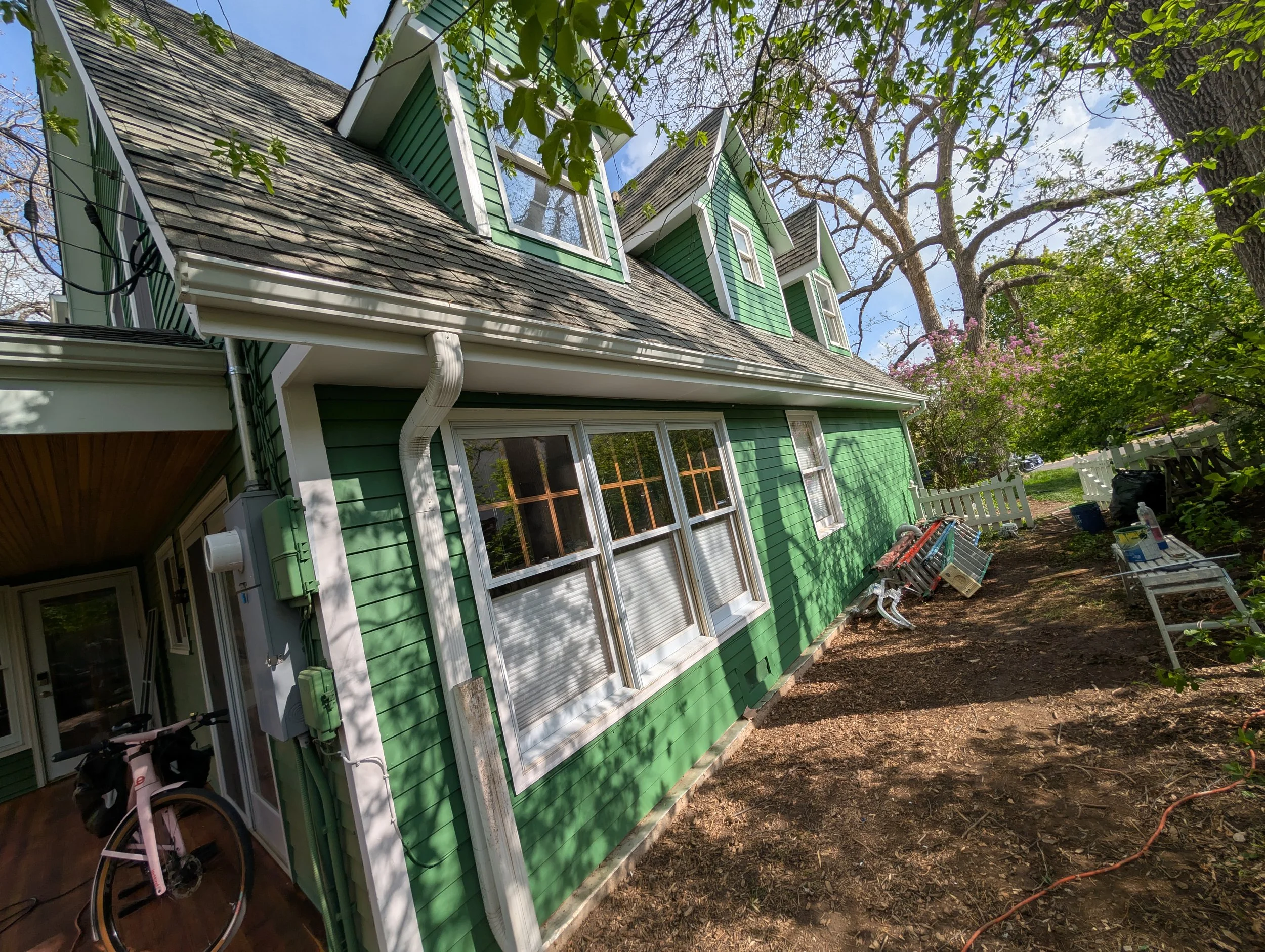 A green house with a sloped roof, multiple dormer windows, and white trim, situated in a yard with trees and outdoor furniture.