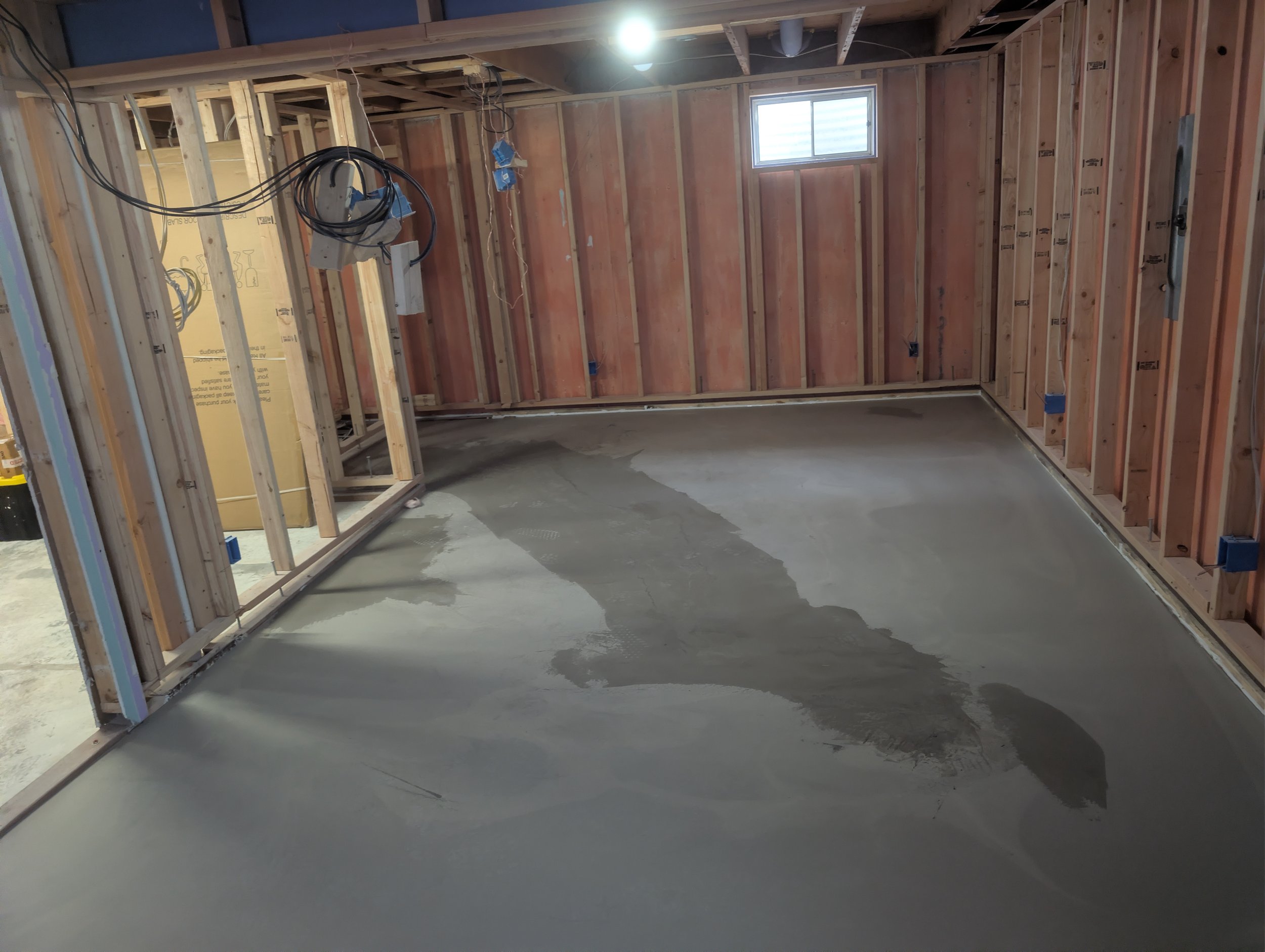 Interior of a house under construction with exposed wooden framing, a small window, electrical wiring, and a freshly poured concrete floor.
