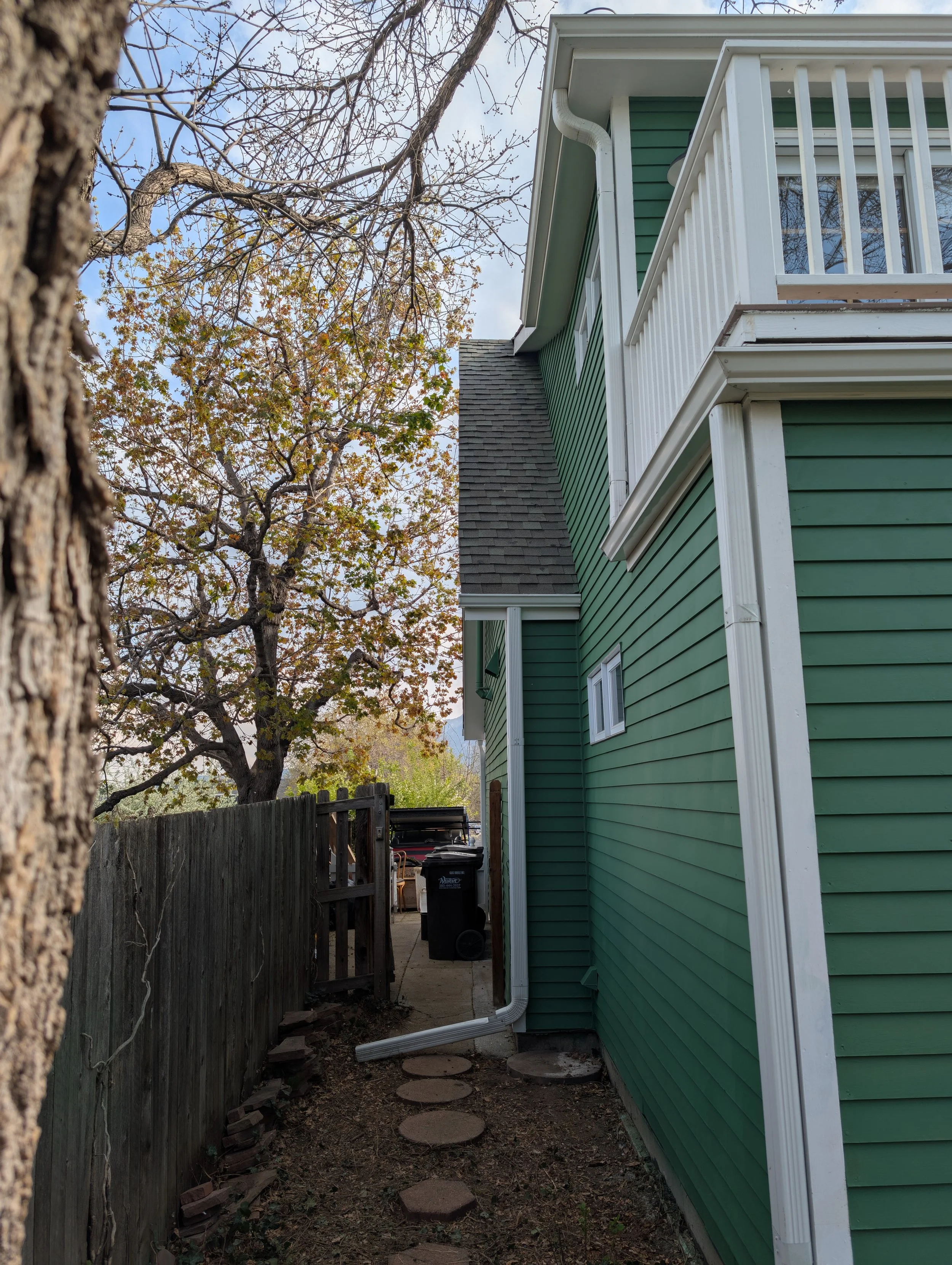 Side yard view of a green house with white trim, a narrow pathway with stepping stones, a tall wooden fence on the left, and trees with autumn leaves.