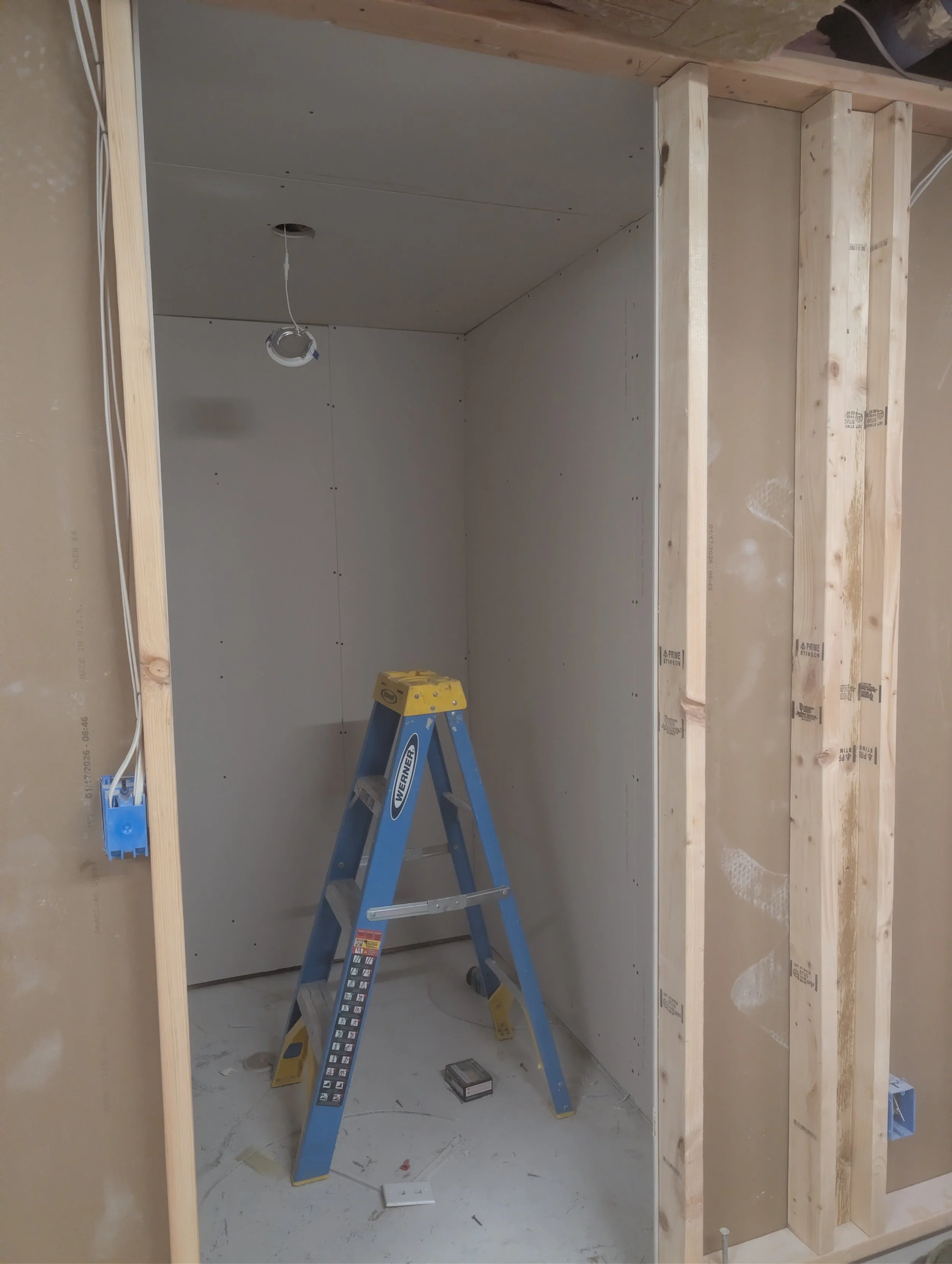 Construction site with wooden framing and drywall being installed, a blue step ladder in the center, and electrical wiring visible.