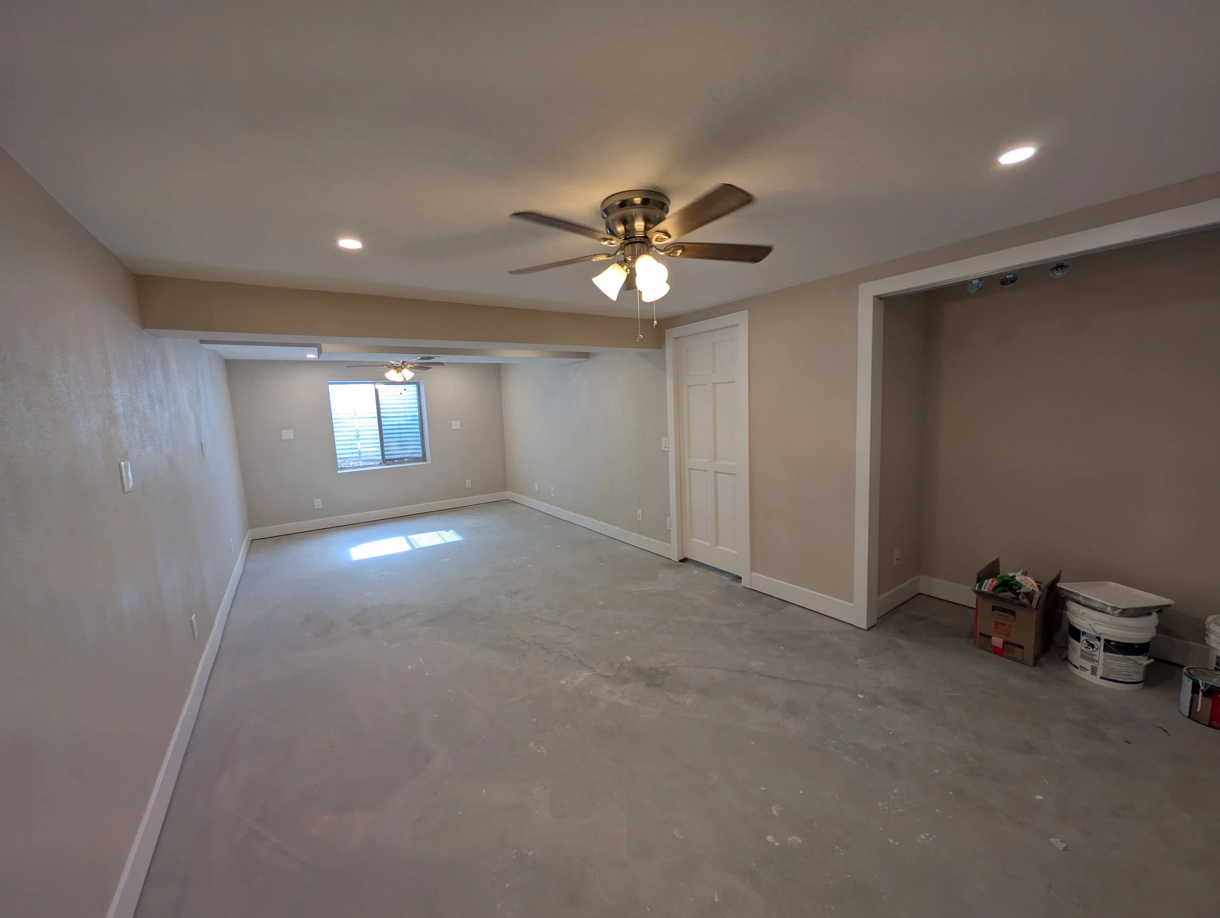 An empty room under renovation with beige walls, a ceiling fan with lights, a window, and paint cans and supplies on the floor.
