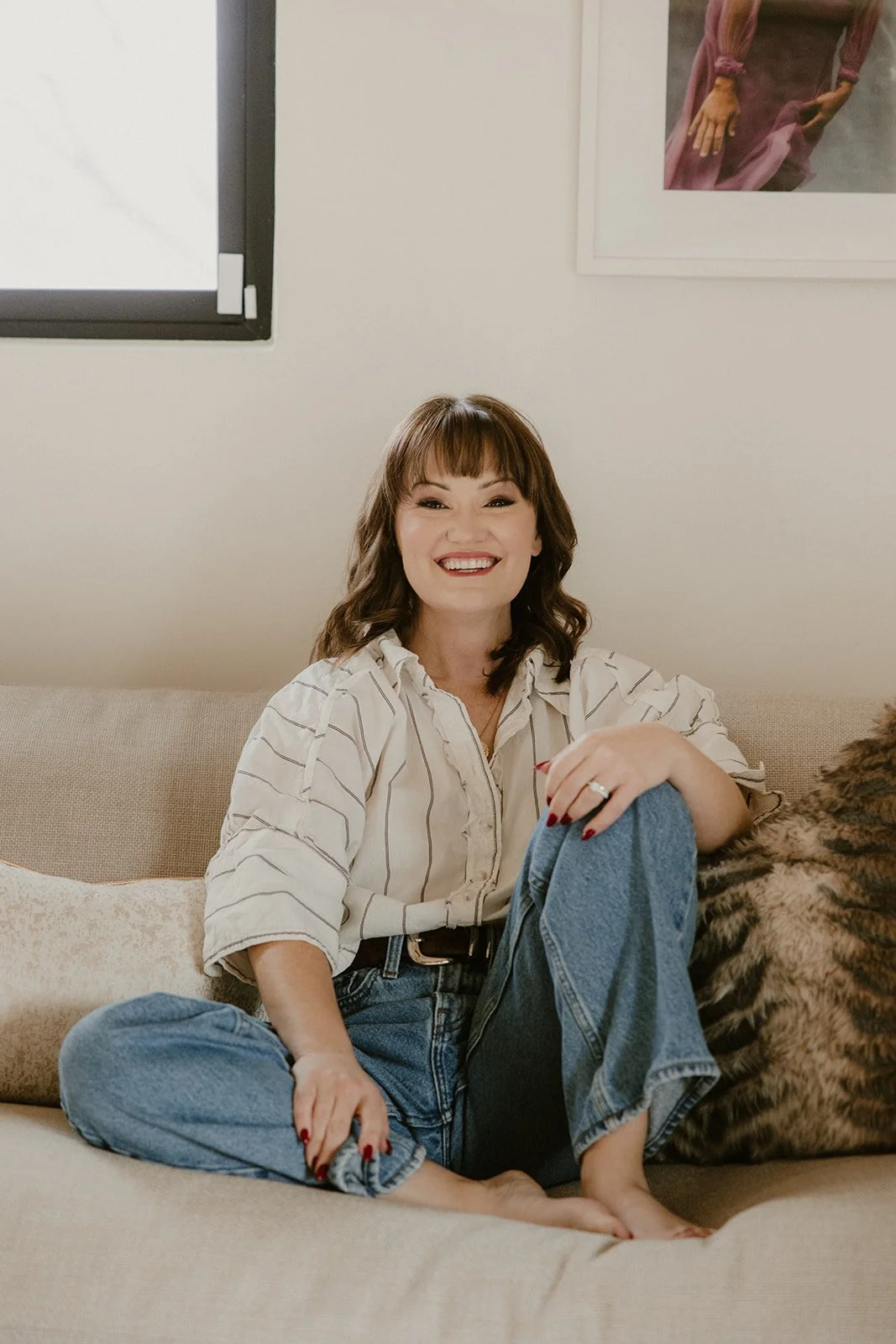 A woman with brown hair sitting cross-legged on a beige sofa, smiling at the camera. She is wearing a cream-colored blouse with vertical stripes and blue jeans.
