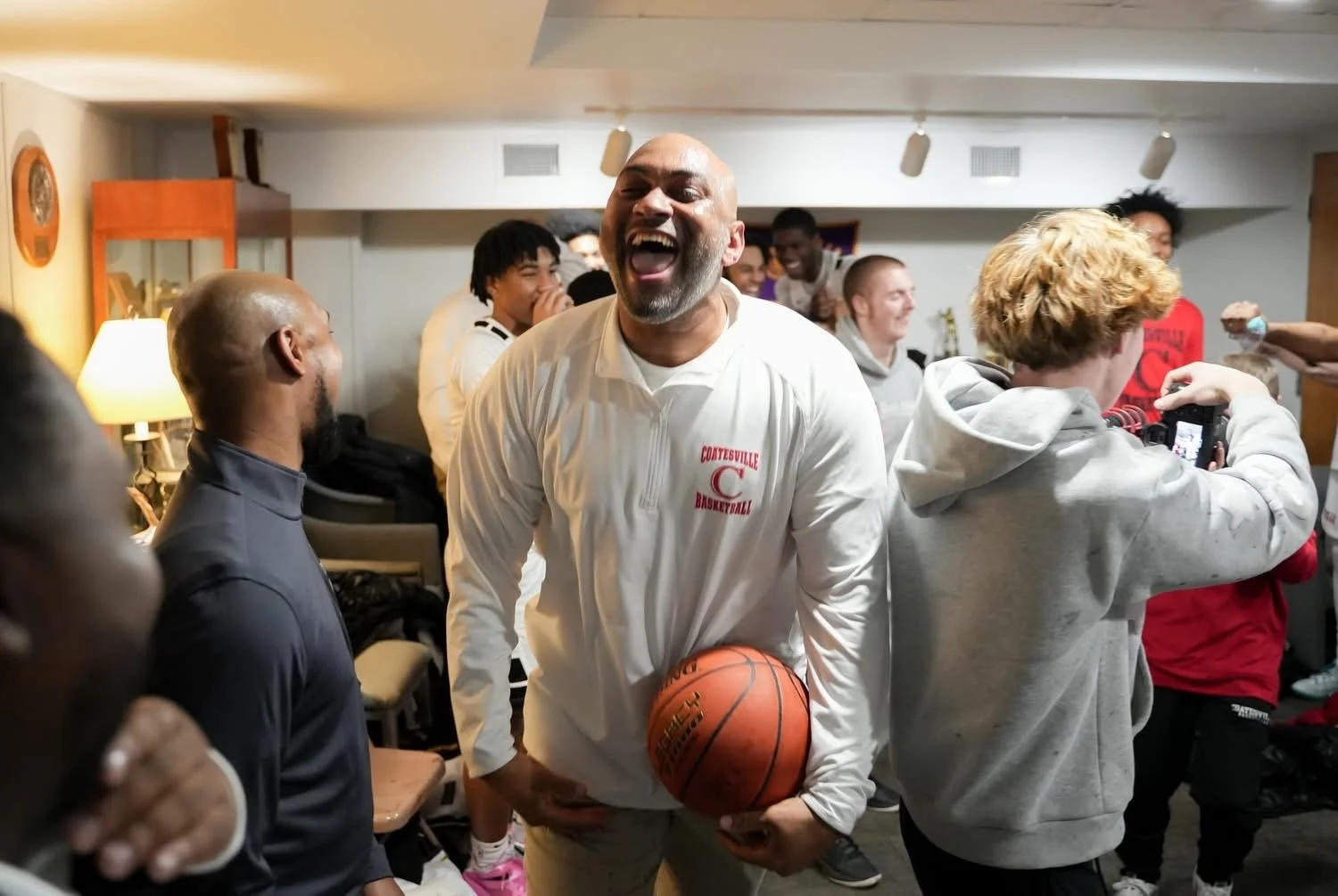 Group of people celebrating in a room, with Coach John Allen smiling holding a basketball in the center. The man is wearing a white shirt with red text, and others around him are laughing and taking photos.
