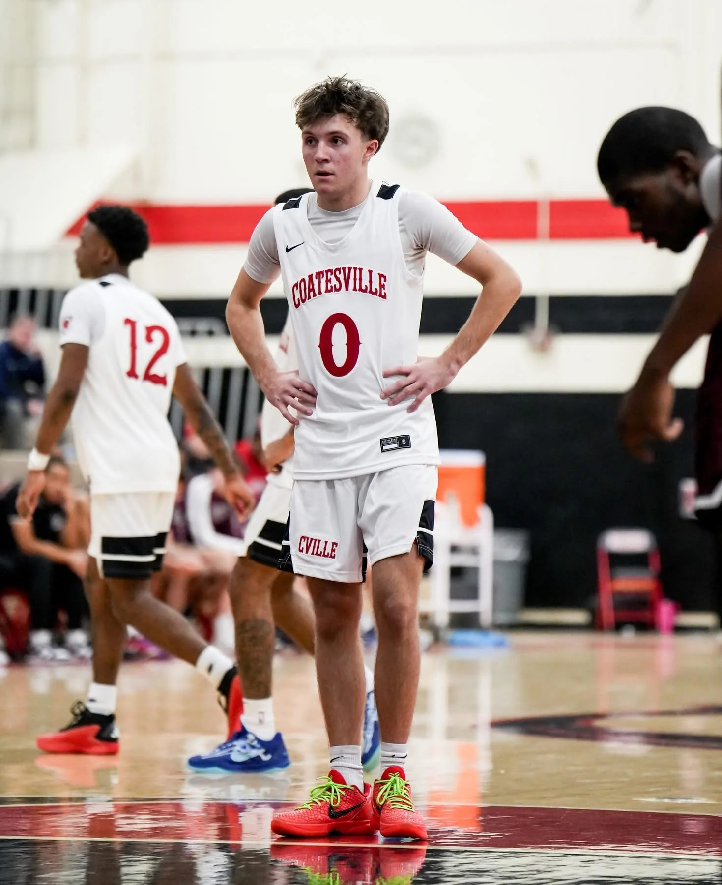 Basketball players on the court during a game, with one player standing with hands on hips, wearing a white jersey with 'COATESVILLE' and the number 0, and others in the background.