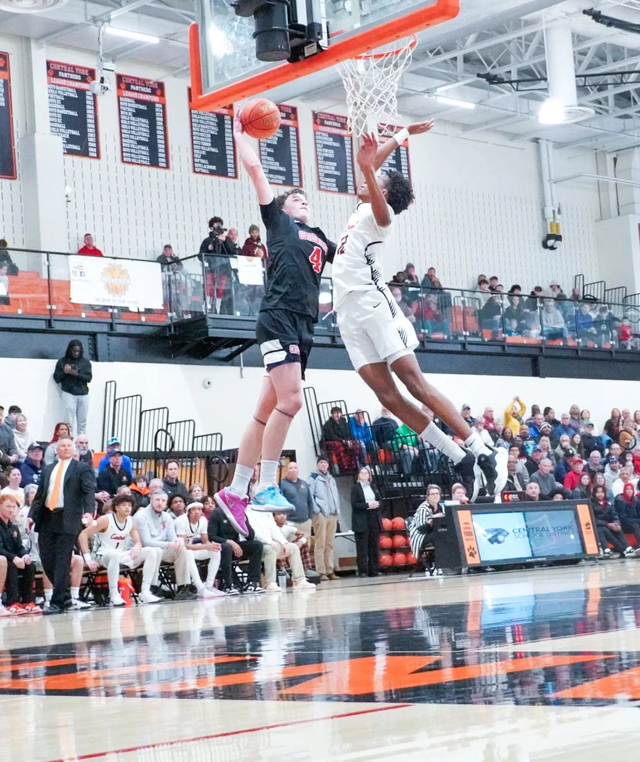 A basketball game with Colton Hiller in black Coatesville jersey number 4 dunking on a player in white uniform. Spectators are sitting and watching in the background.