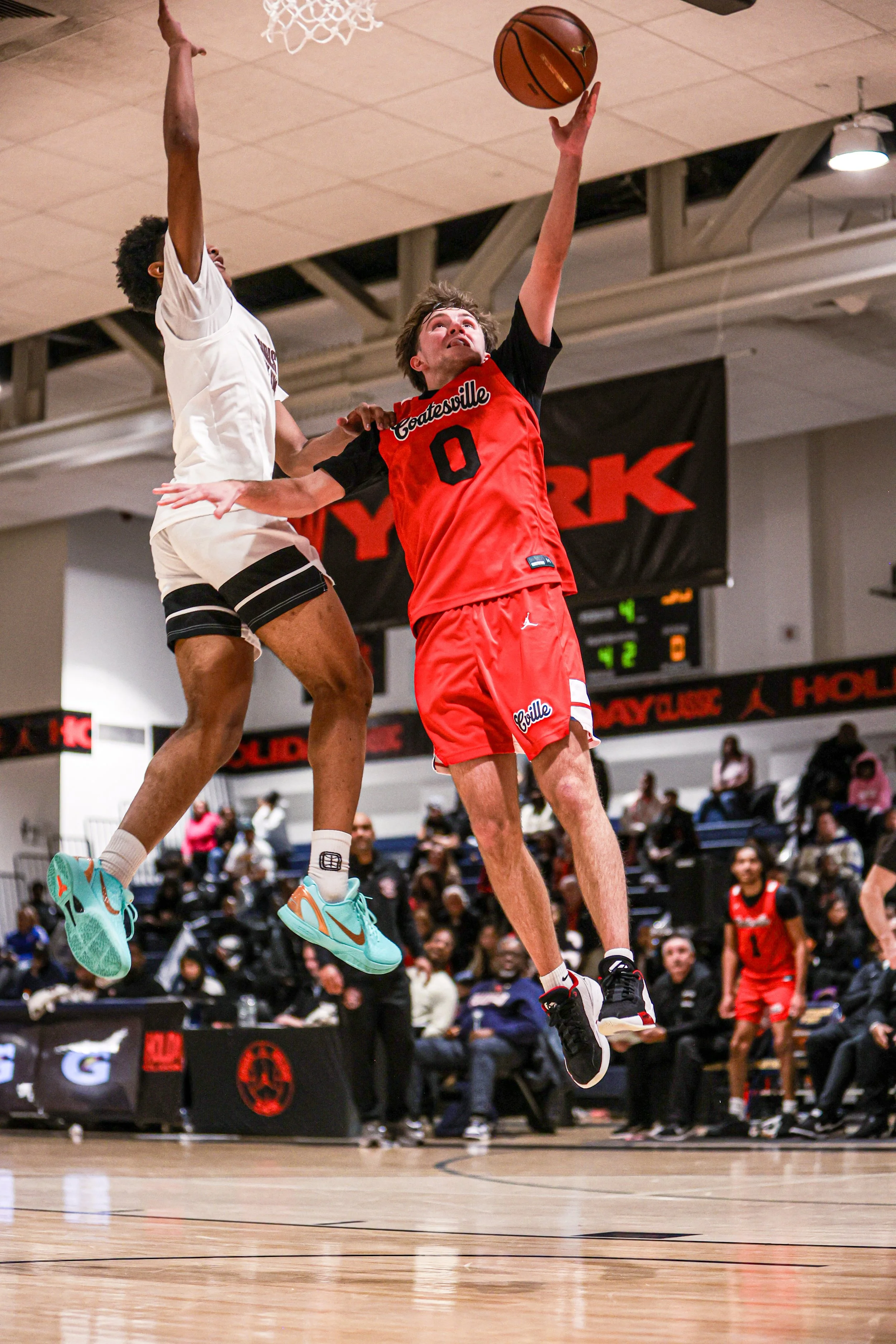 Group of people celebrating in a room, with Coach John Allen smiling holding a basketball in the center. The man is wearing a white shirt with red text, and others around him are laughing and taking photos.