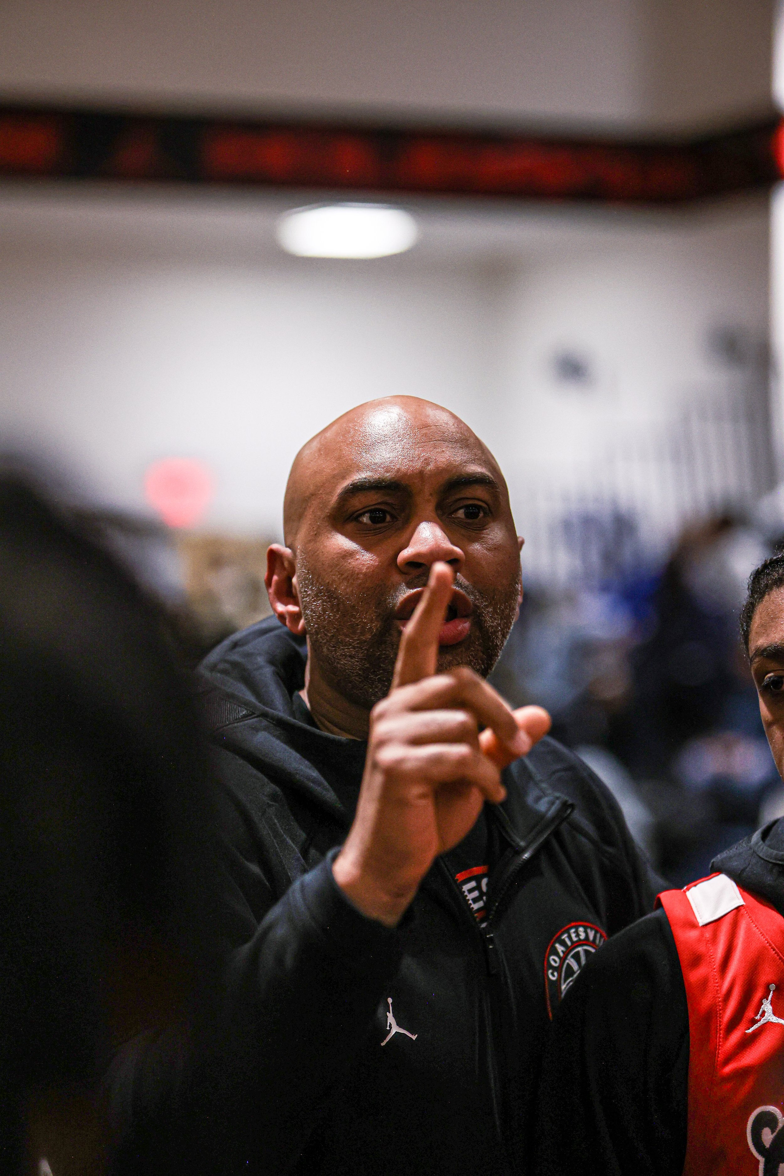 Basketball players on the court during a game, with one player standing with hands on hips, wearing a white jersey with 'COATESVILLE' and the number 0, and others in the background.