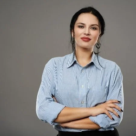 A woman with dark hair pulled back, wearing a blue and white striped button-up shirt, standing with arms crossed against a neutral gray background.