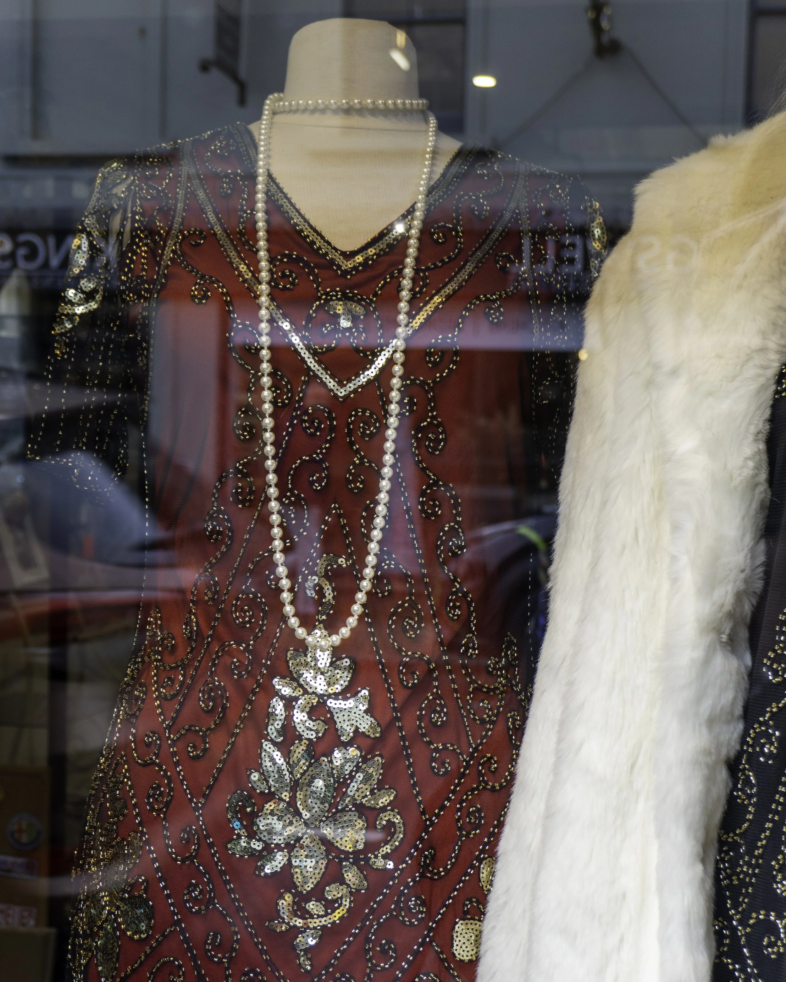 A mannequin dressed in a red and black sequined dress with intricate gold and black embroidery, wearing a long pearl necklace. Part of a white fur coat is visible on the right side.