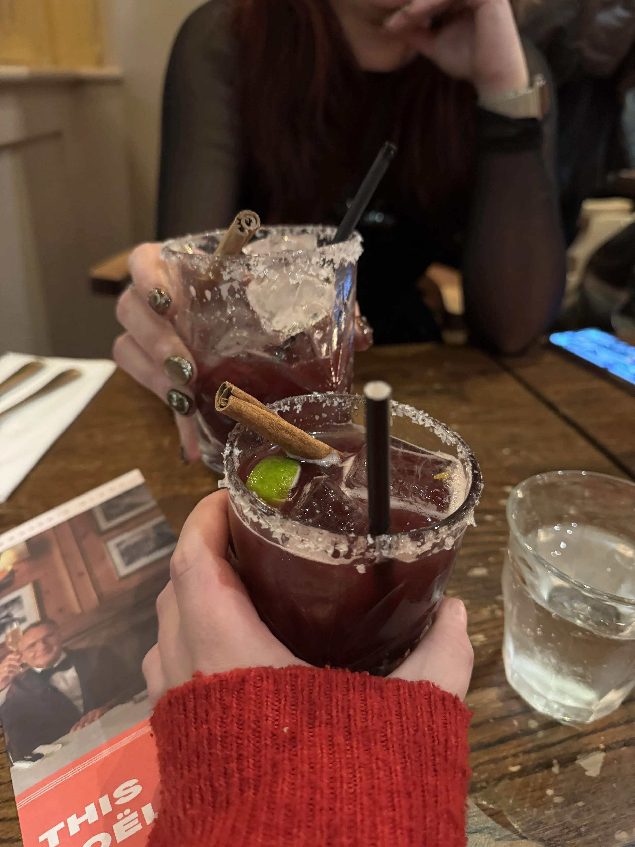 Two people holding glasses of dark red cocktails with salt-rimmed edges, garnished with cinnamon sticks and lime wedges, at a restaurant table.