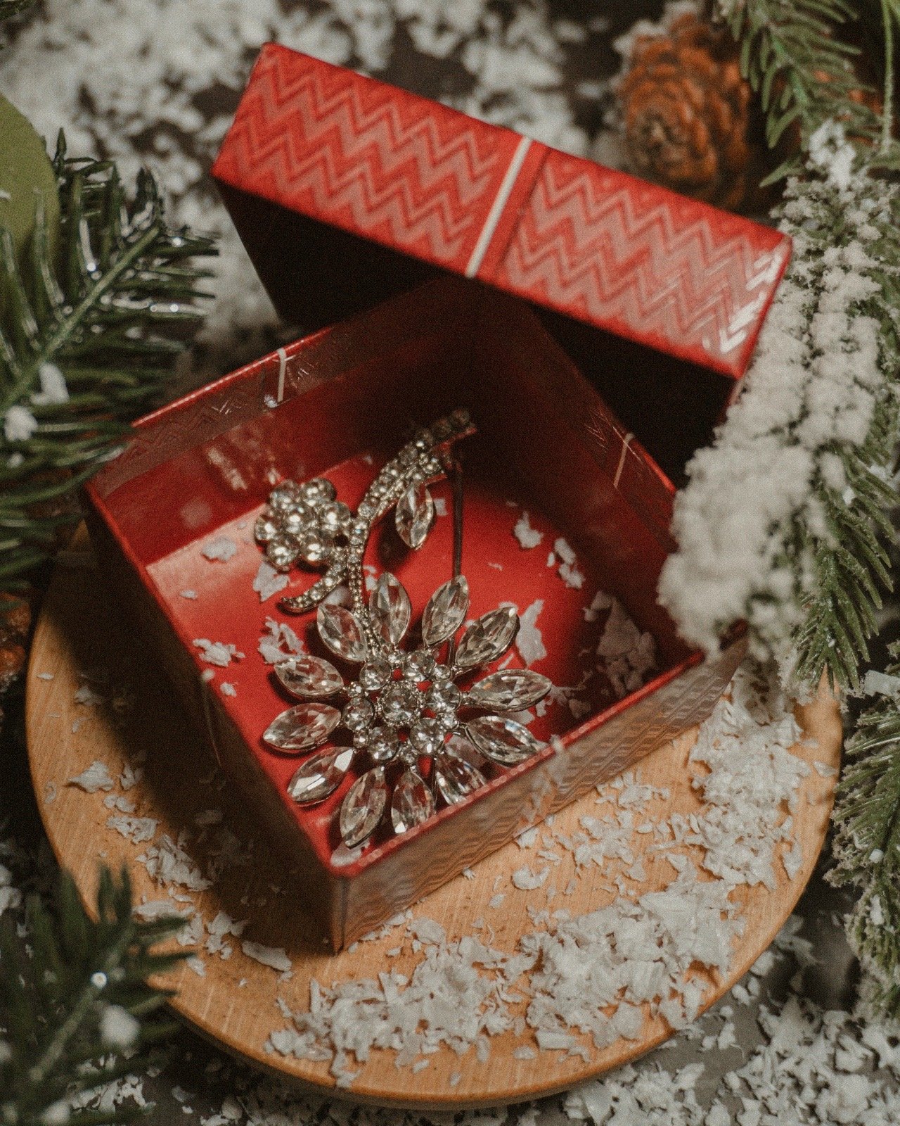 A jewelry box with a pair of sparkly earrings inside, surrounded by Christmas decorations including snow, pine branches, and a pine cone.