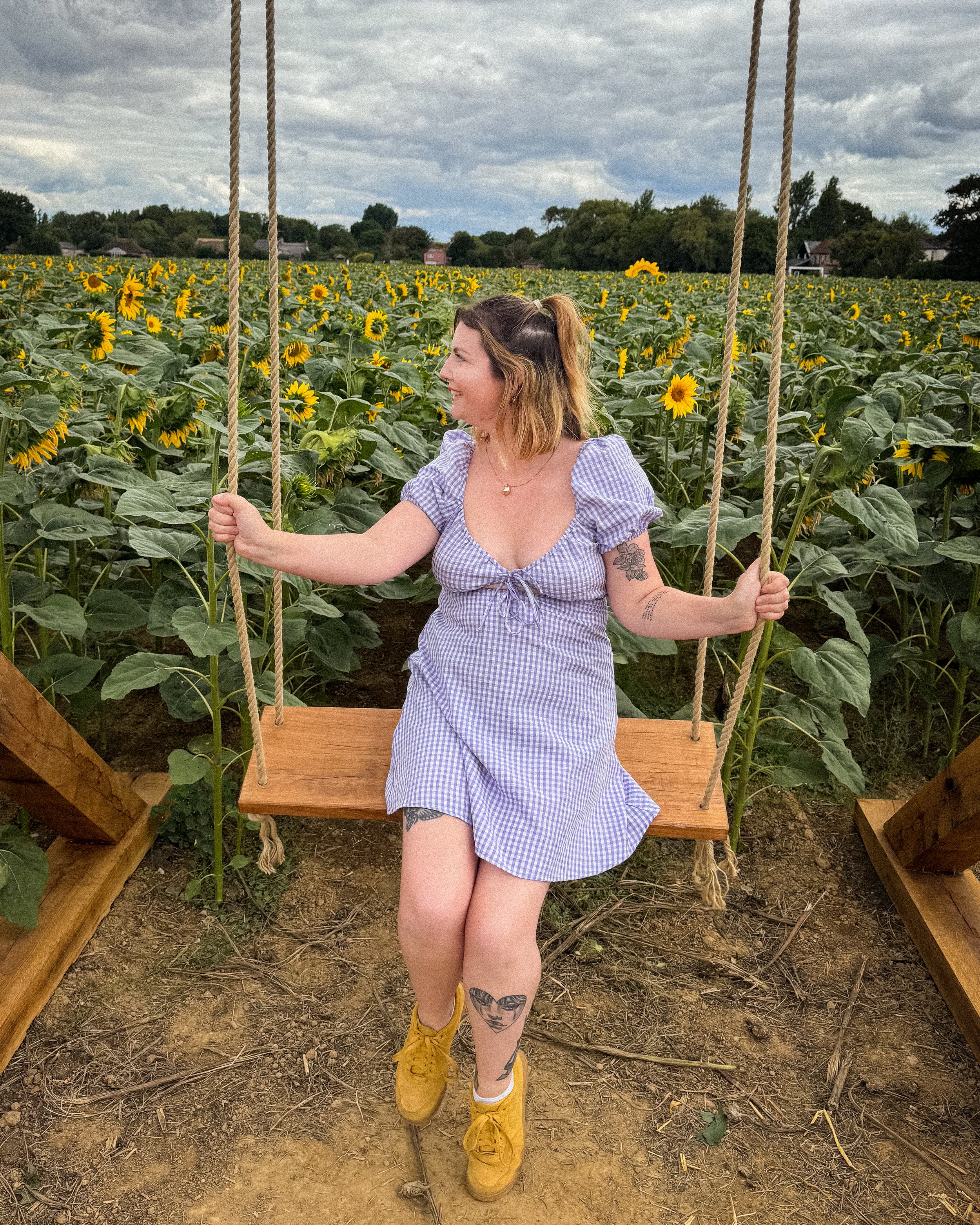 A woman with tattoos and shoulder-length hair sitting on a wooden swing in a sunflower field, wearing a blue and white gingham dress and yellow boots.