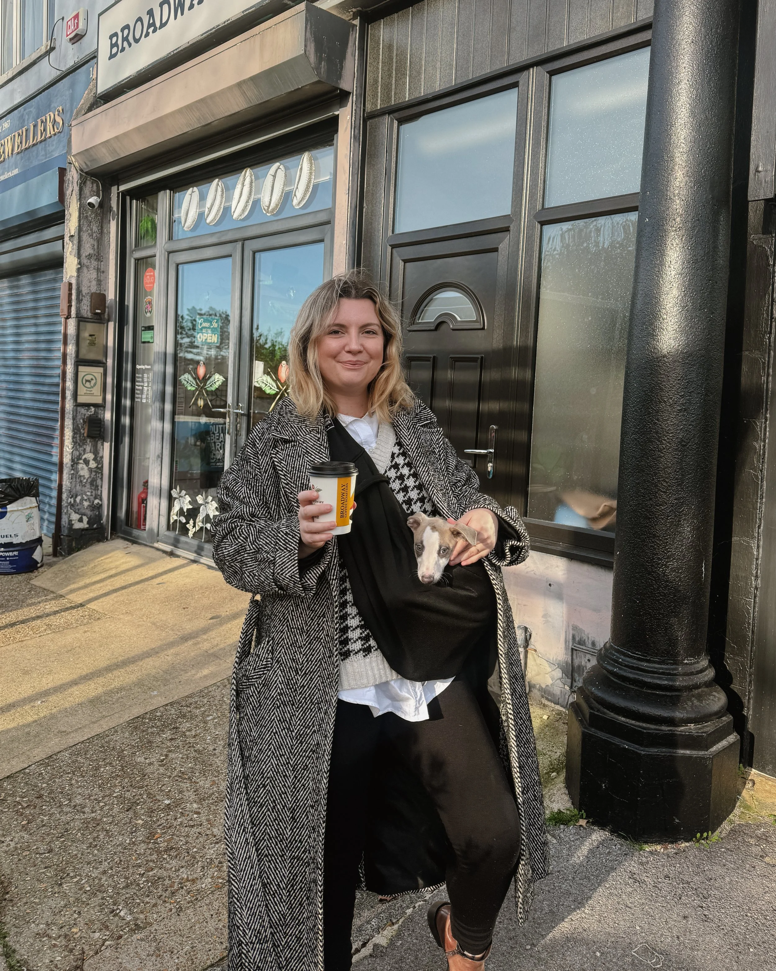 A woman with shoulder-length blonde hair smiling while holding a coffee cup and a small dog in a black carrier bag, standing outside a storefront with a sign that says 'BROADWAY.'