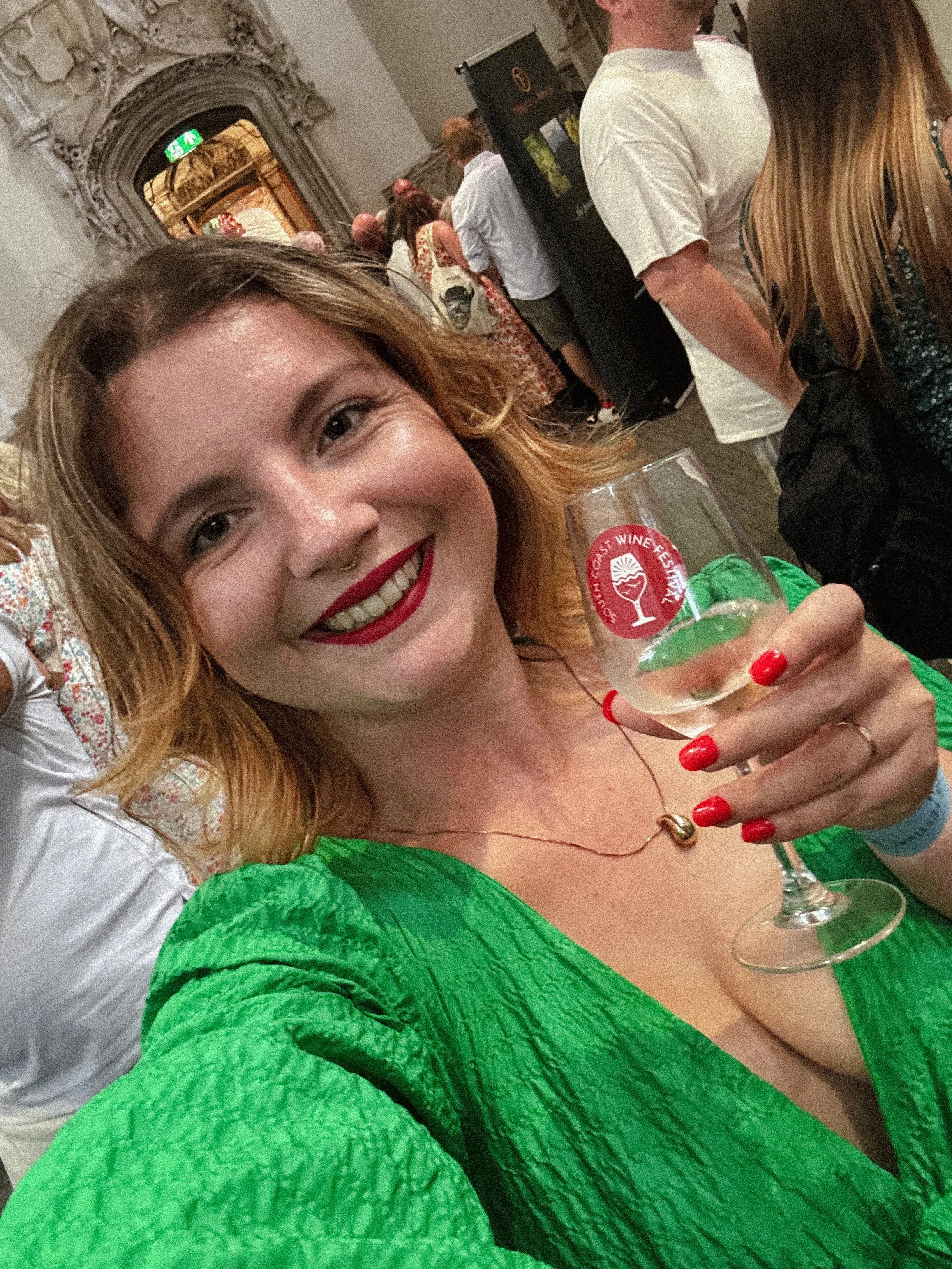 A smiling woman with red hair and red lipstick, wearing a green dress and holding a glass of wine at a wine festival, with other people in the background.